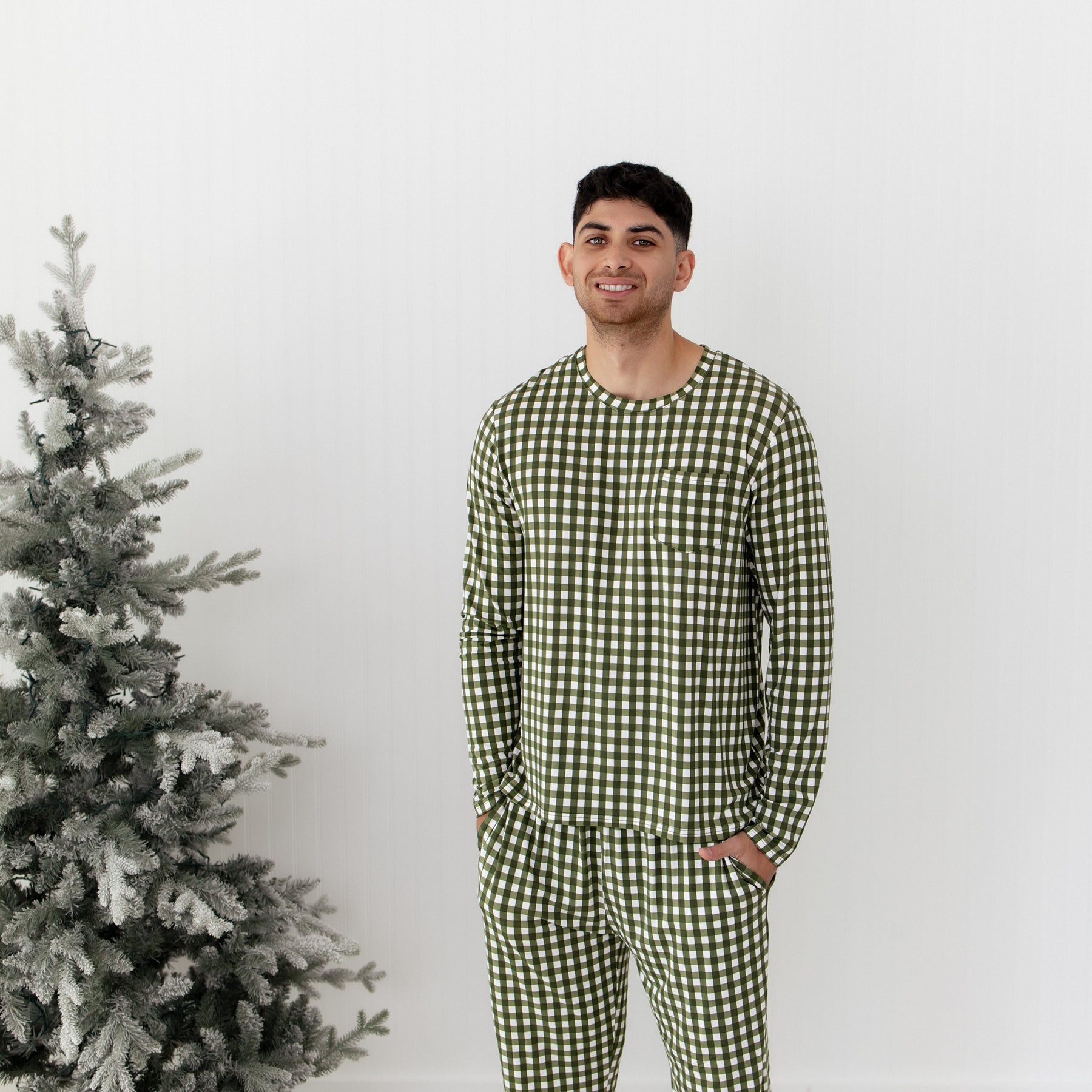 Man wearing green and white checkered pajamas standing next to a decorated Christmas tree on a plain background