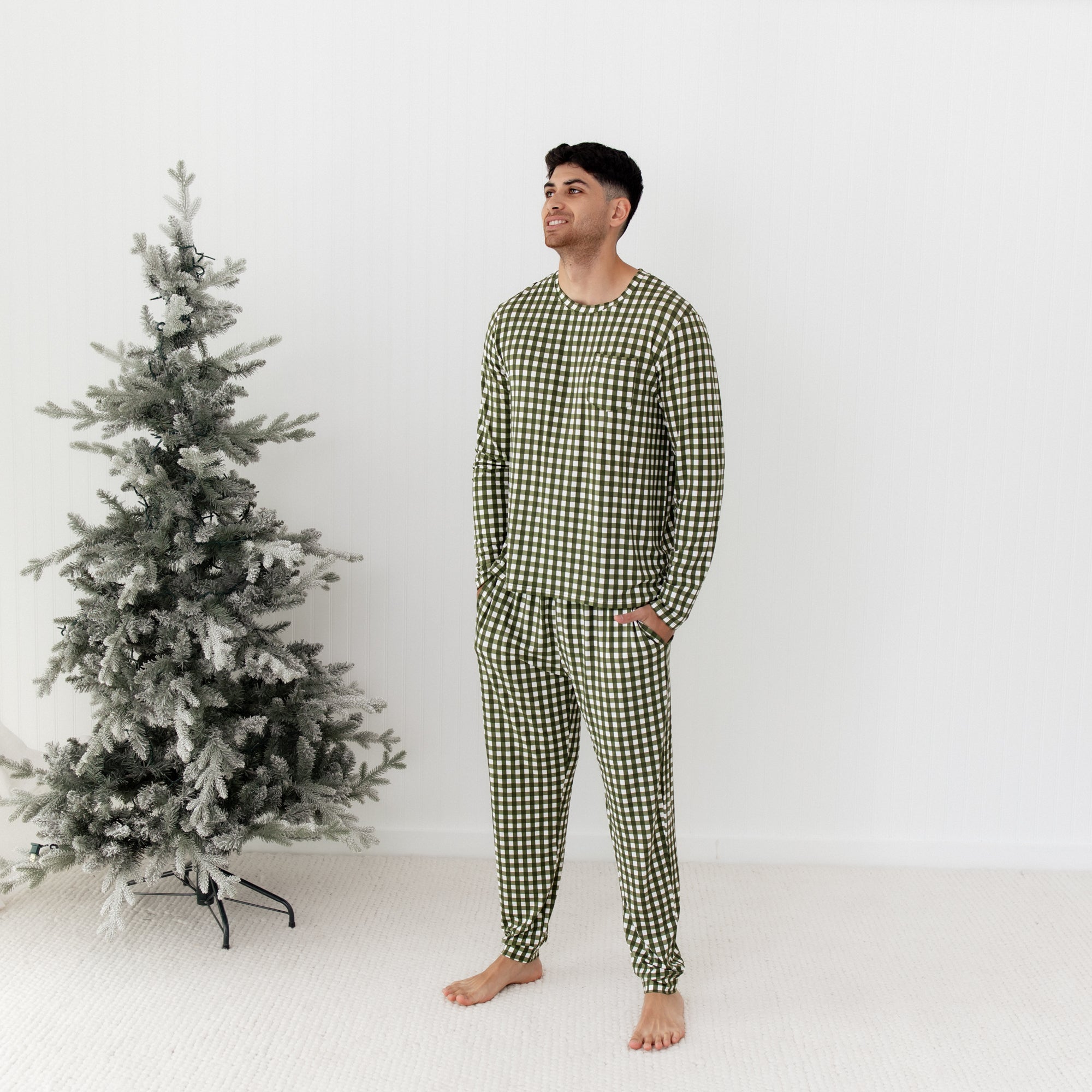 Man wearing green checkered pajamas standing next to a decorated Christmas tree on a white background
