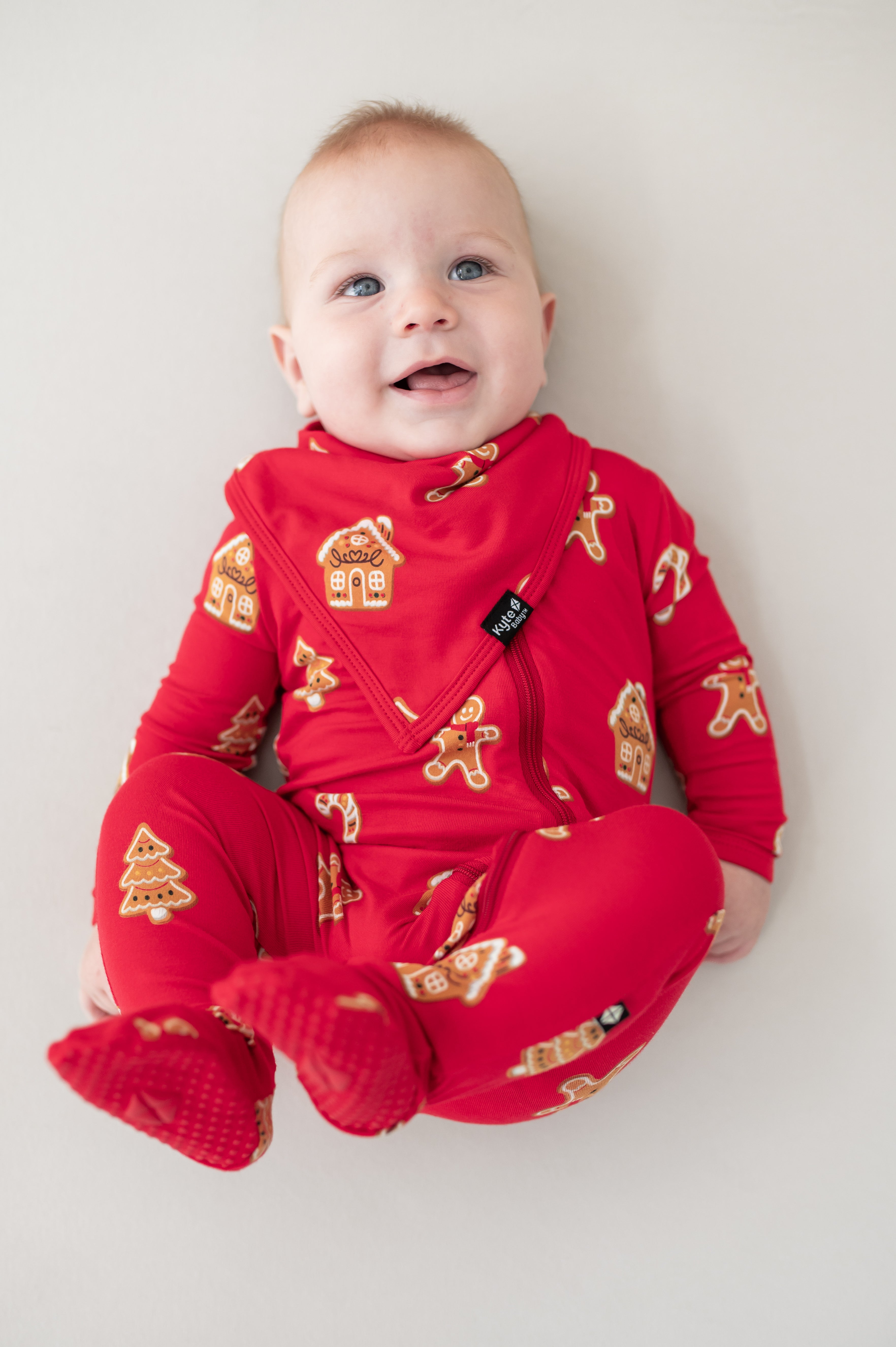 Baby wearing Bib in Gingerbread print over similar print zippered footie lying on a beige background