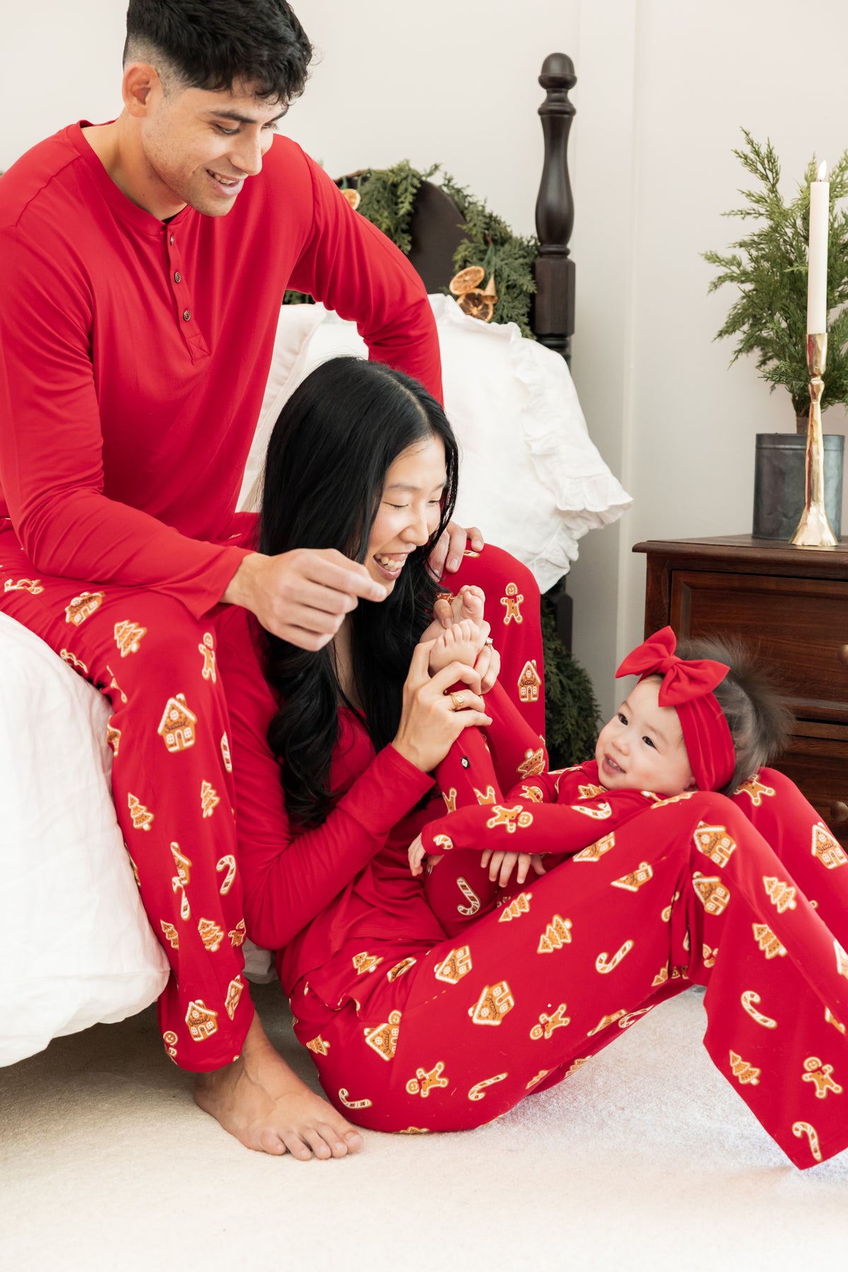 A family of three wearing matching red holiday pajamas with gingerbread patterns, sitting together on a white carpet in a bedroom setting.