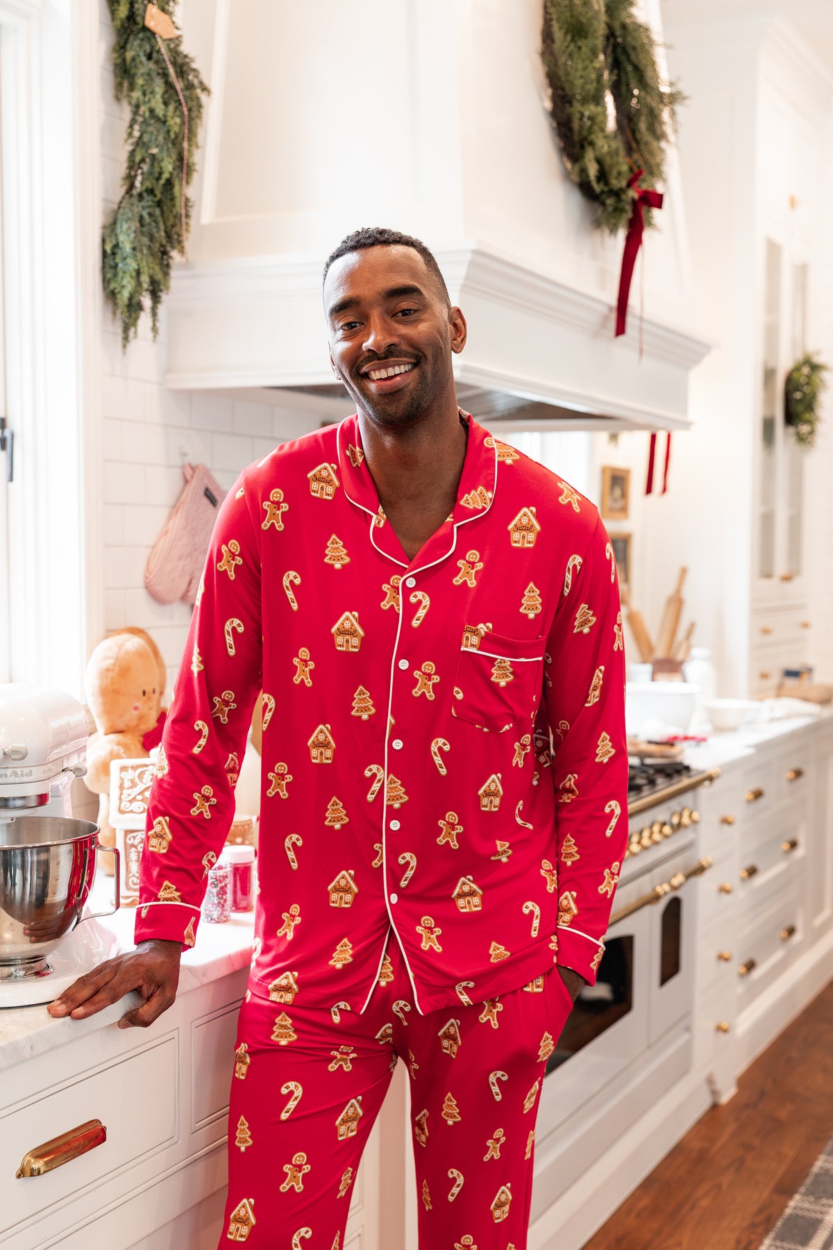 A man wearing a festive red and gold holiday-themed pajama set stands in a bright, white kitchen decorated with wreaths.