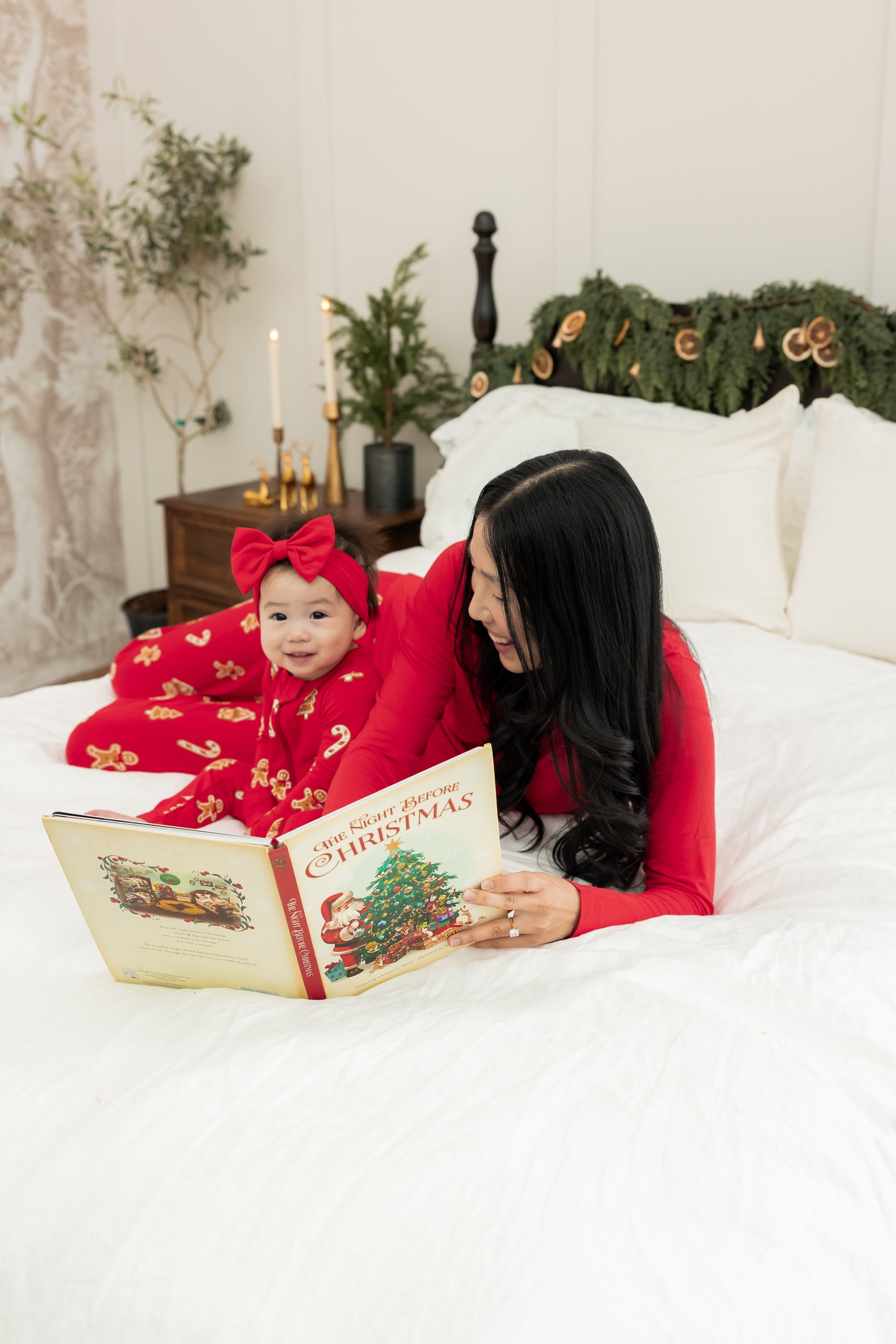 A woman and a toddler lie on a white bed reading a Christmas book together. The woman wears a bright red long-sleeve top, and the toddler wears matching red pajamas with a red bow headband.