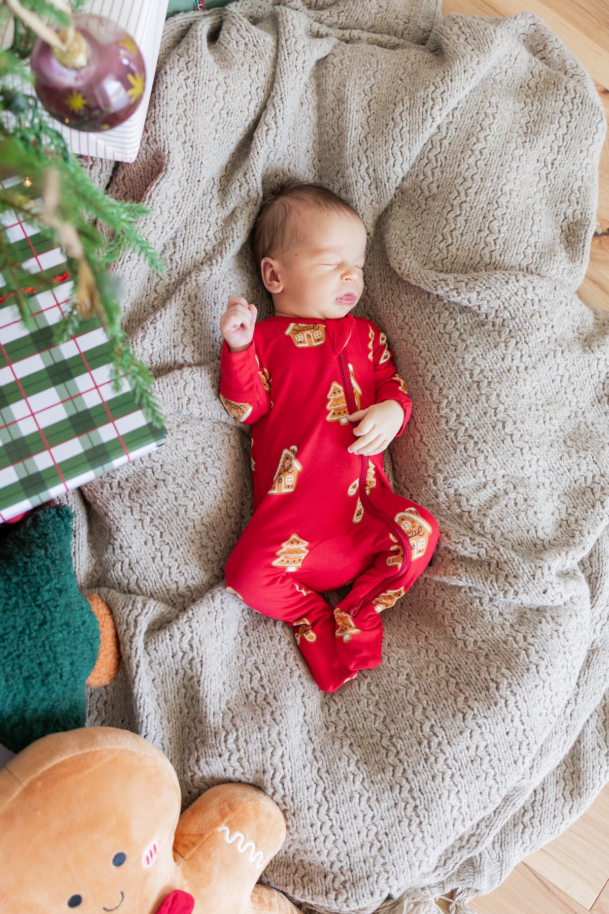 A sleeping baby wearing a red footed onesie with a gold Christmas tree pattern, lying on a textured gray blanket with holiday decorations in the background.