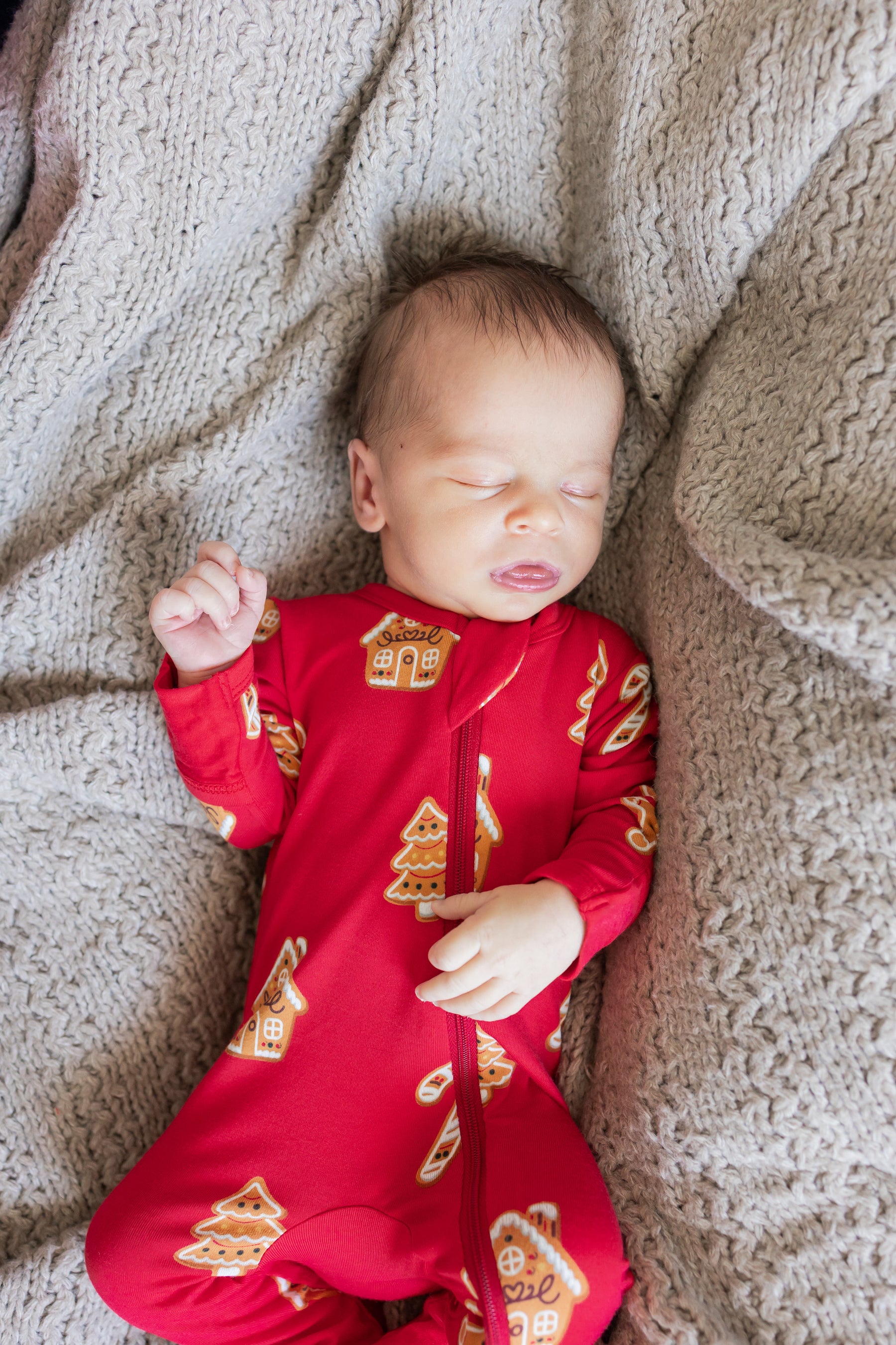 A sleeping newborn baby wearing a red one-piece pajama with gingerbread house and tree patterns, lying on a textured beige knit blanket.