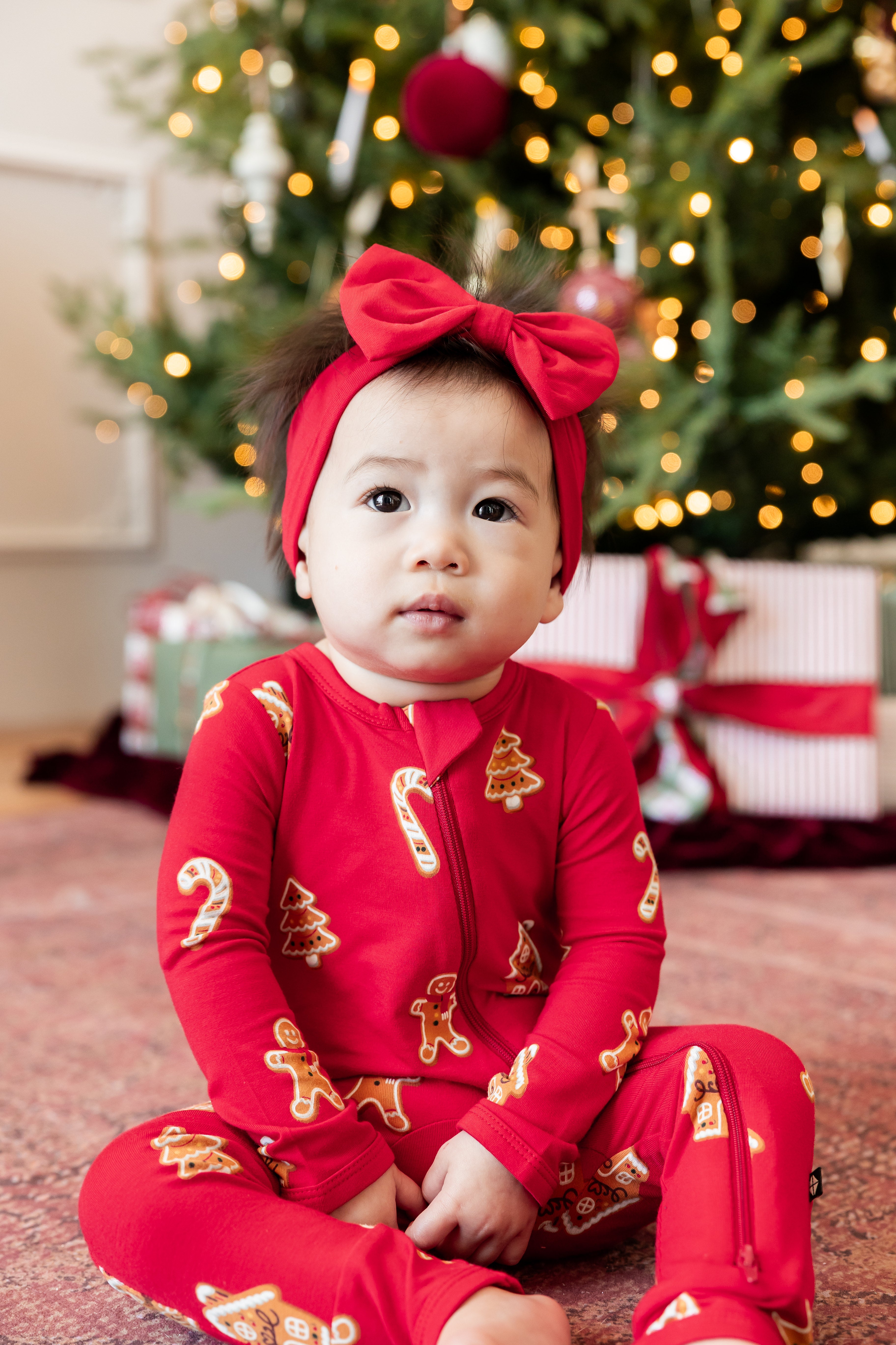A baby sitting on a carpet in front of a decorated Christmas tree, wearing a red zip-up one-piece pajama with gingerbread man and candy cane prints, along with a matching red bow headband.