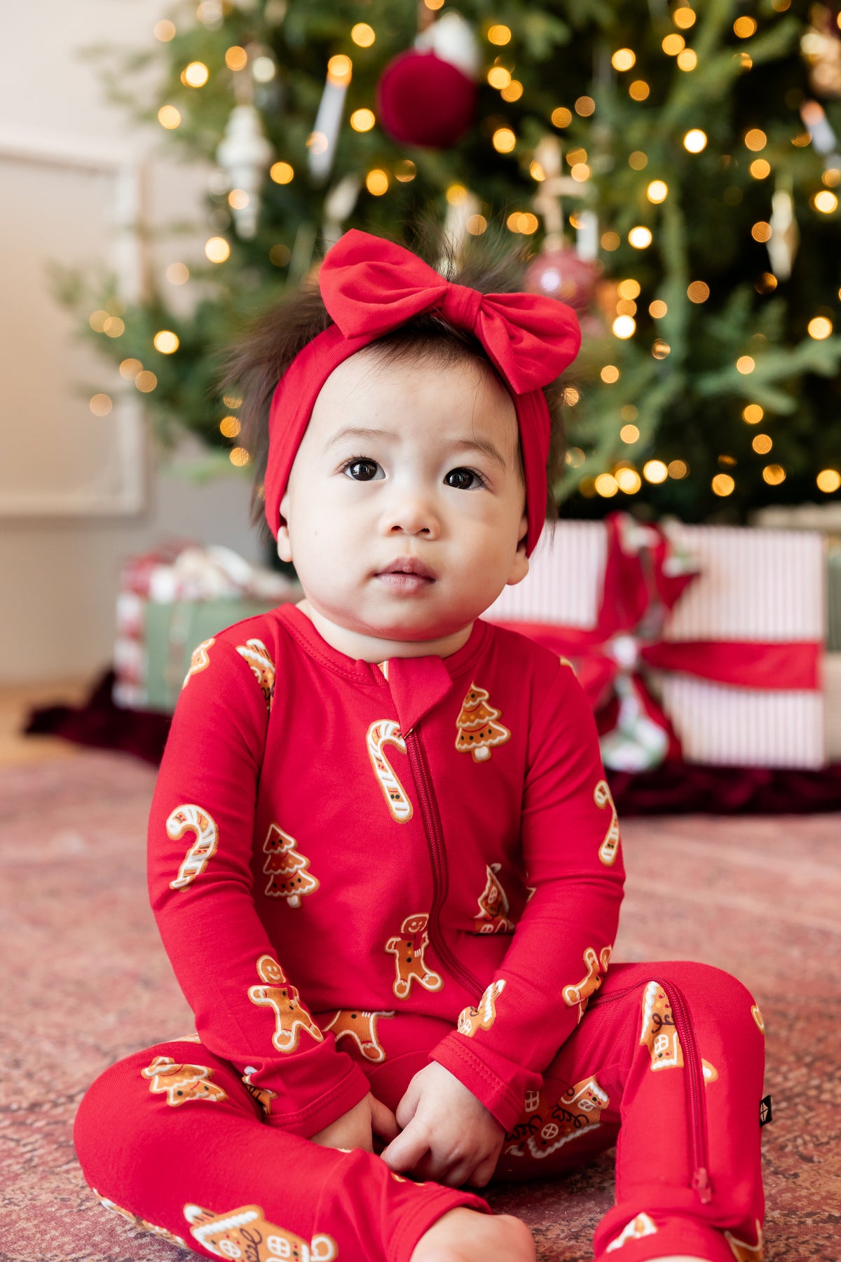 A baby sitting on a carpet in front of a decorated Christmas tree, wearing a red zip-up one-piece pajama with gingerbread man and candy cane prints, along with a matching red bow headband.