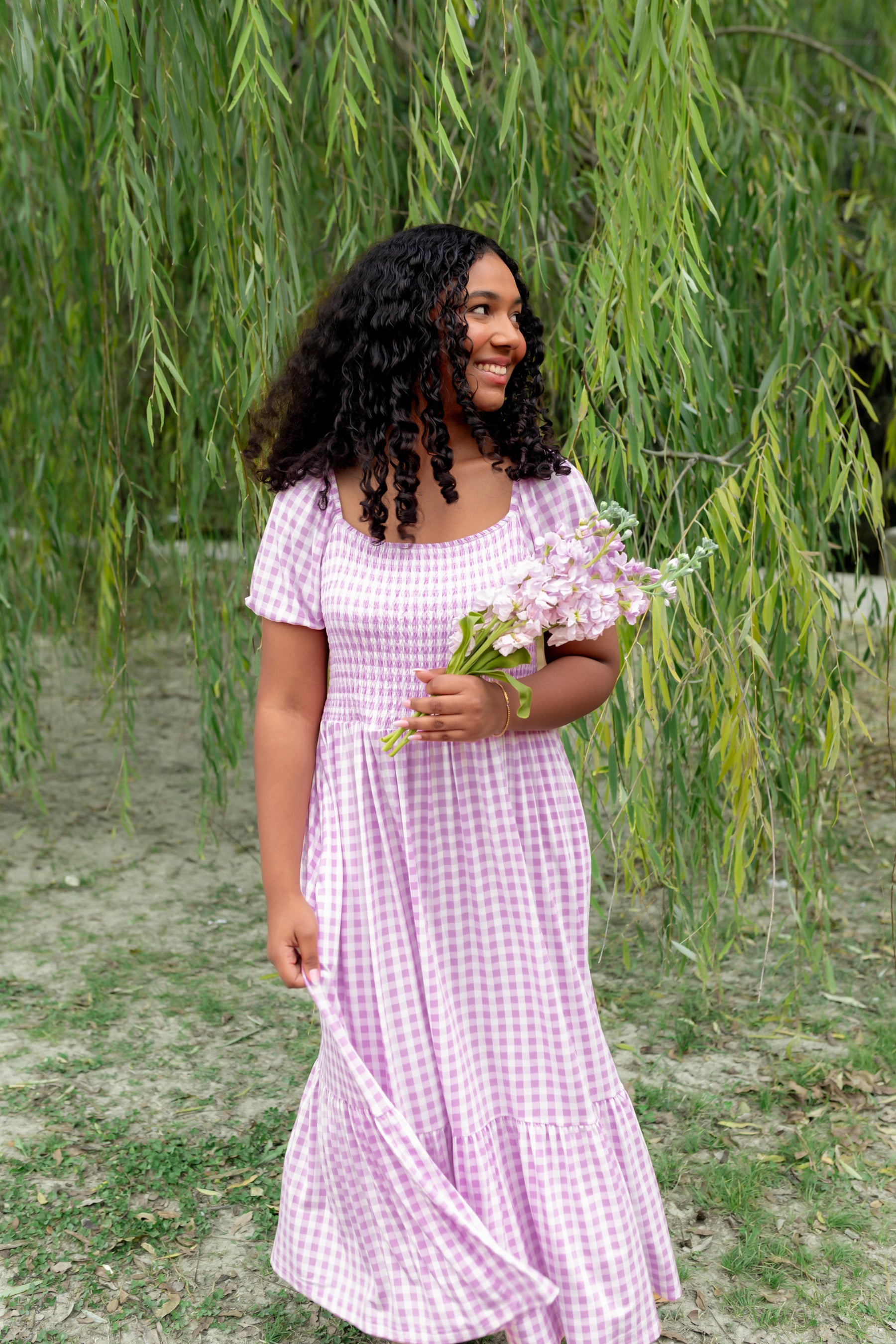 Smiling model standing in a field in front of a tree wearing the Women’s Smocked Dress in Gingham Thistle holding light purple flowers