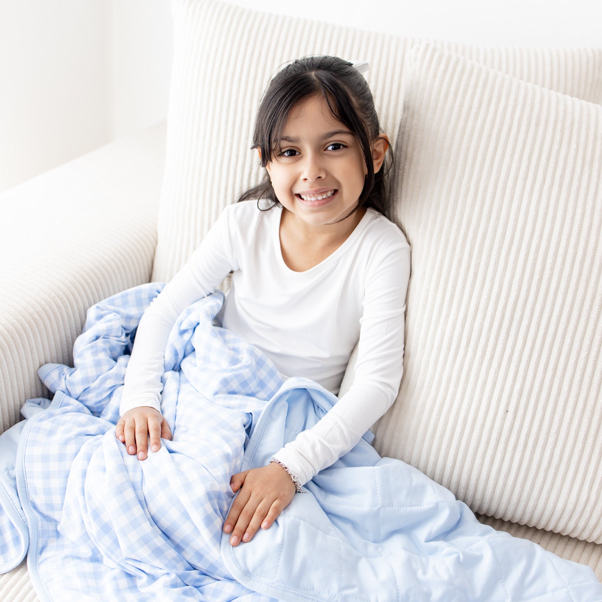 Young girl sitting on a cream couch wearing white long sleeve pajamas with the Toddler Blanket in Gingham Breeze 1.0 over her lap