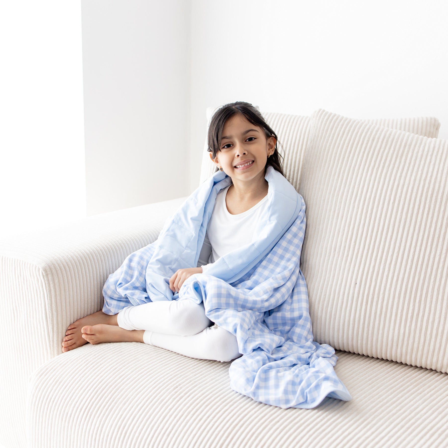 Young girl sitting on a cream colored couch wearing white long sleeve pajamas with the Toddler Blanket in Gingham Breeze 1.0 around her shoulders