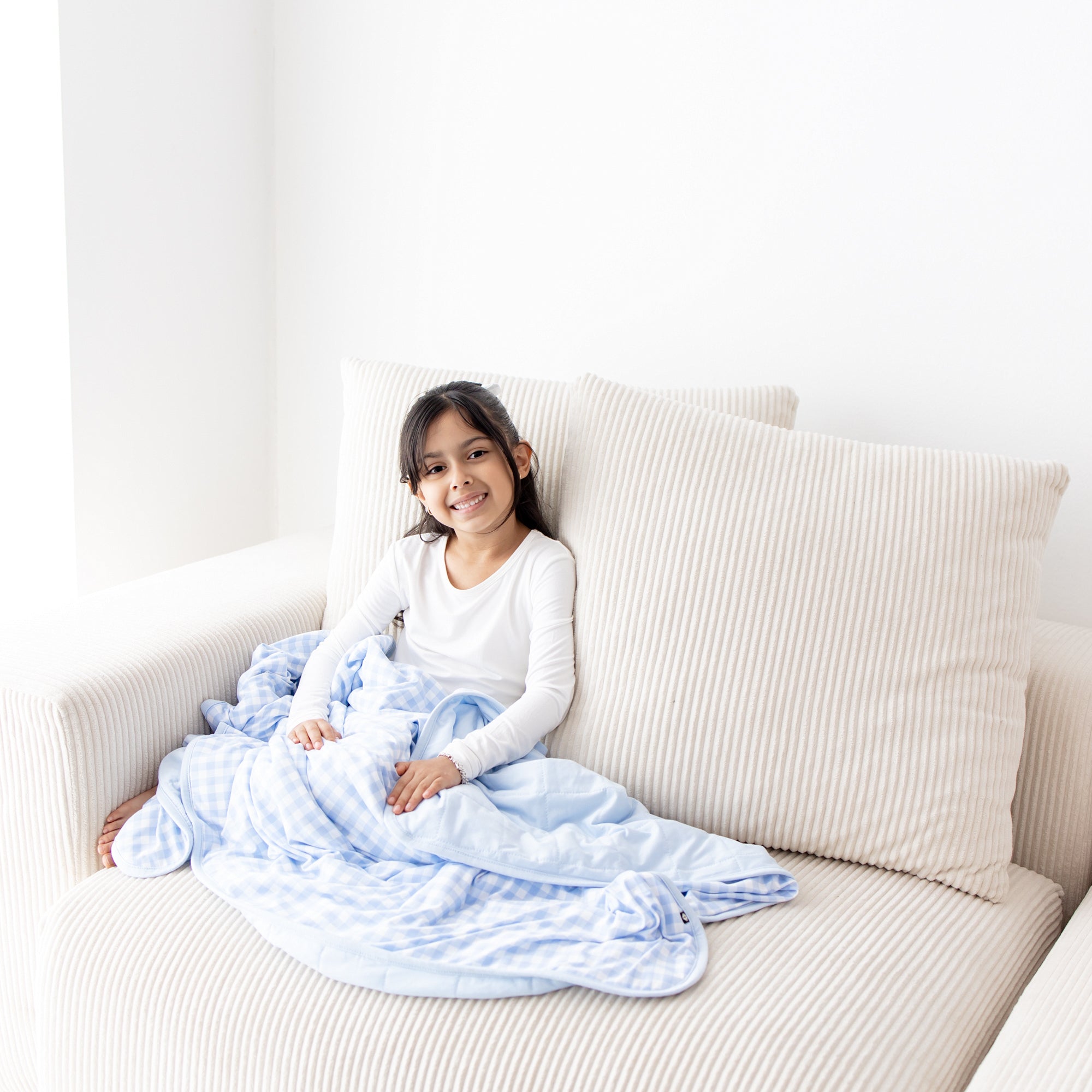 Young smiling girl sitting in the corner of a white cream couch wearing white long sleeve pajamas with the Toddler Blanket in Gingham Breeze 1.0 over her lap