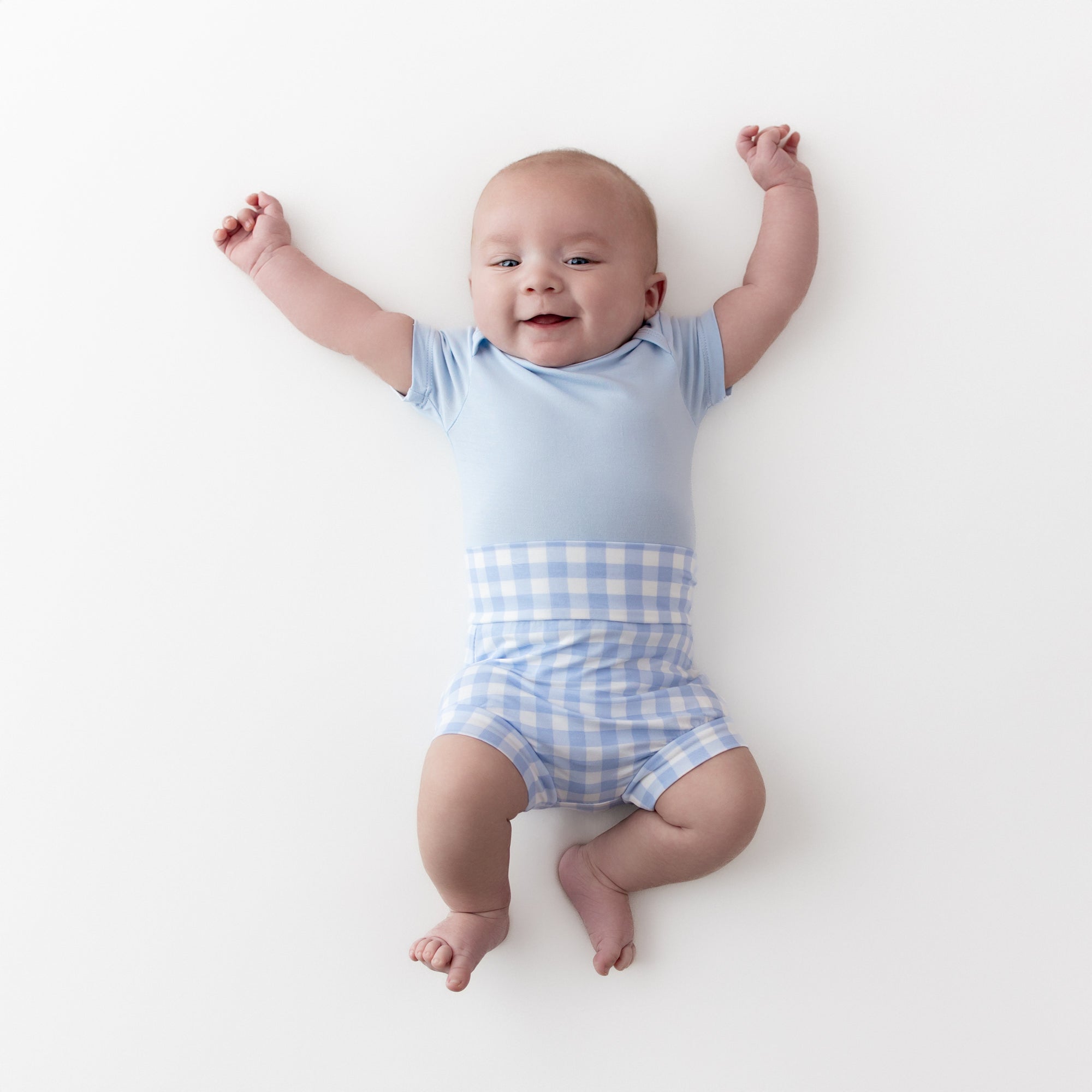 Young infant stretching out wearing the Bummies in Gingham Breeze and light blue short sleeve bodysuit