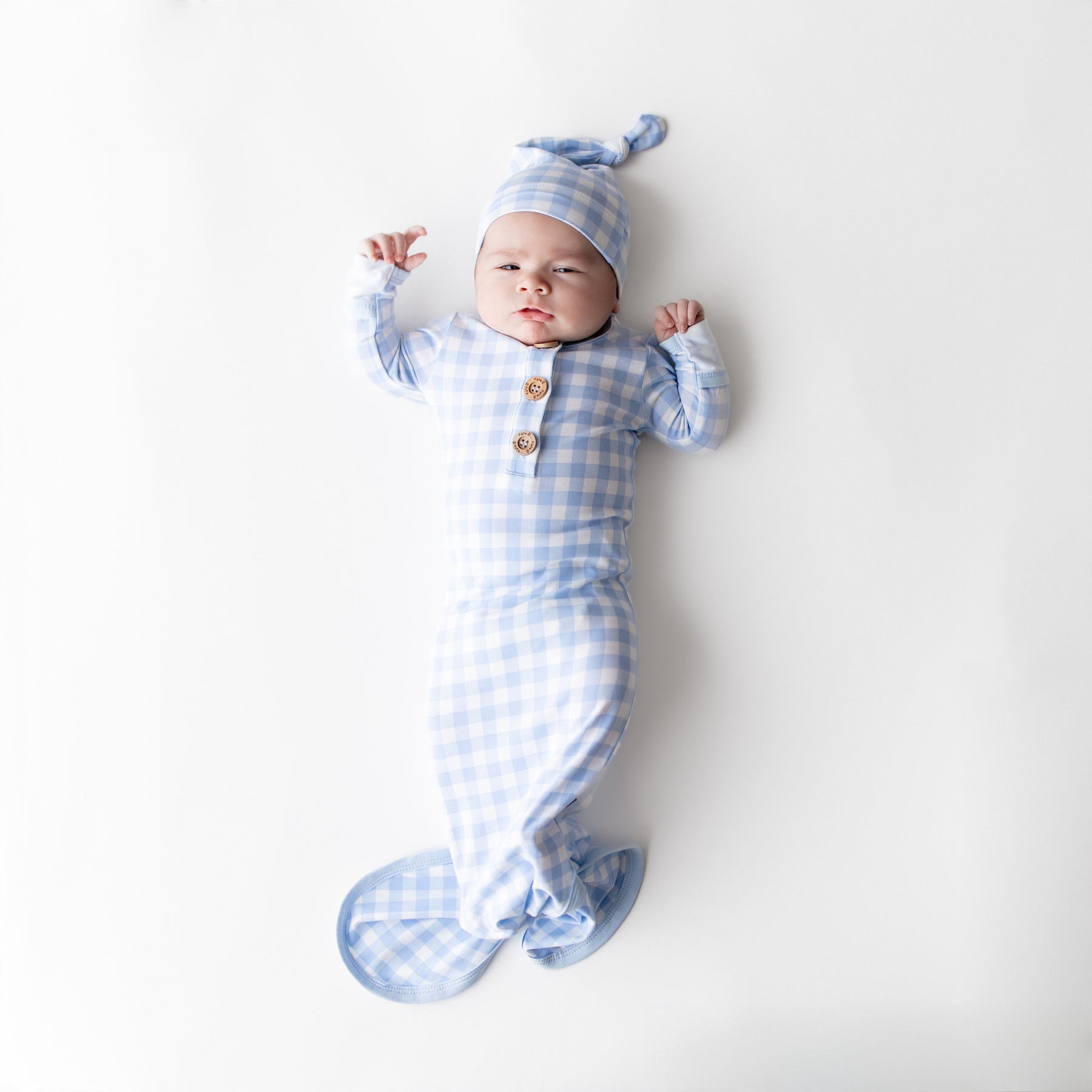Newborn wearing the Knotted Gown with Hat Set in Gingham Breeze laying on a light cream surface