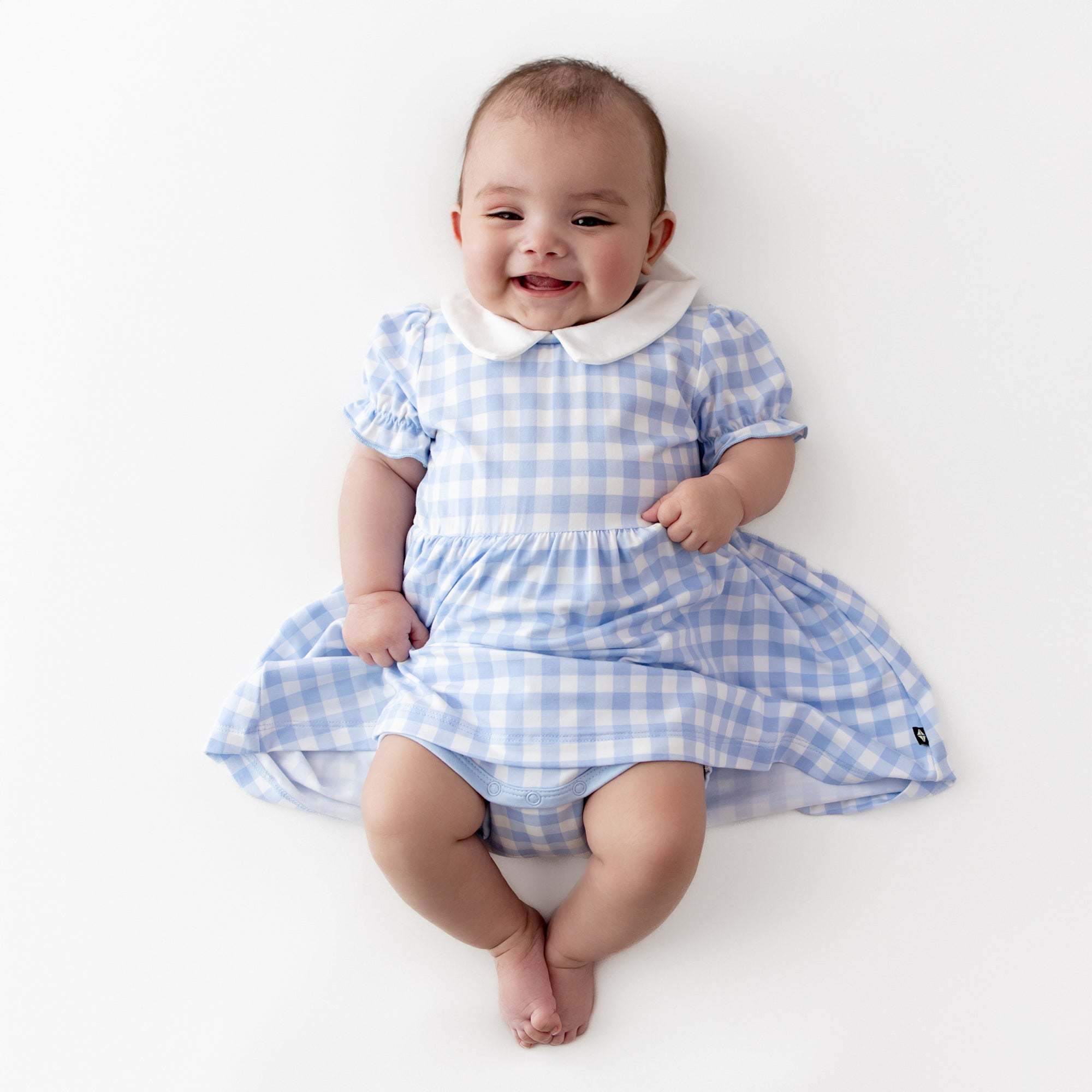 Infant girl wearing the Peter Pan Collar Bodysuit Dress in Gingham Breeze laying on a light cream surface