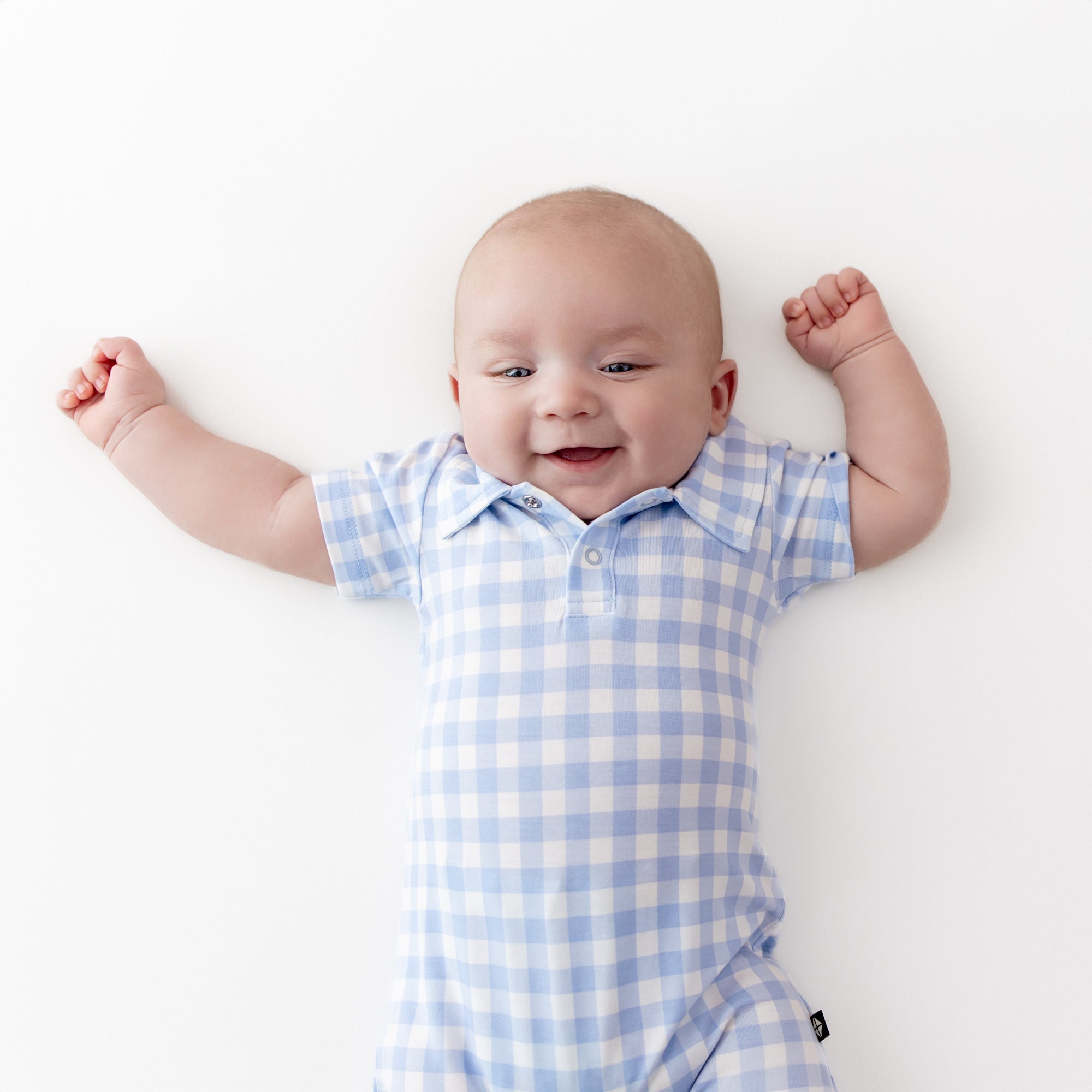 Close up of Polo Shortall in Gingham Breeze shown on an infant laying on a light cream surface