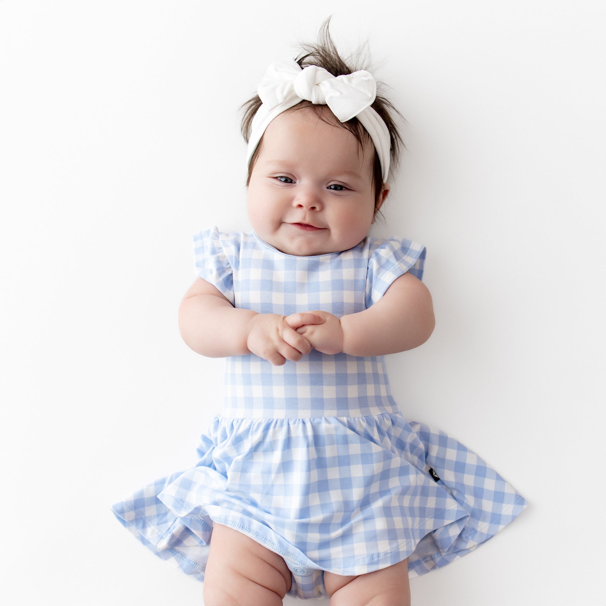 Infant laying on a light cream surface wearing the Twirl Bodysuit Dress in Gingham Breeze with a white knotted bow headband