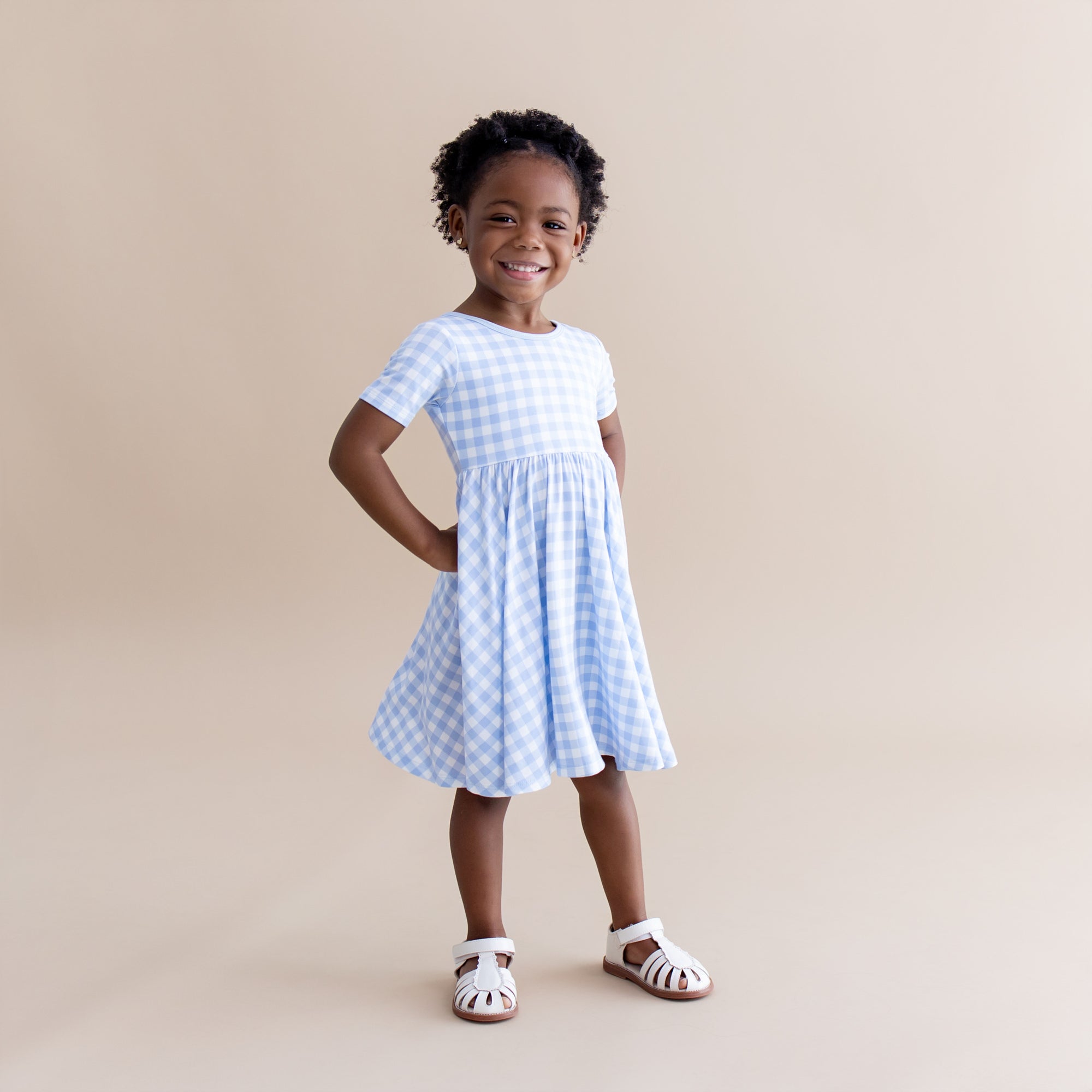 Young girl posing wearing the Twirl Dress in Gingham Breeze paired with white sandals