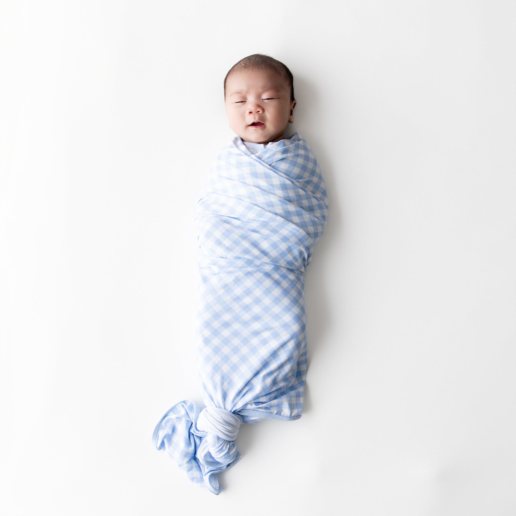 Infant laying on a white surface swaddled in the Swaddle Blanket in Gingham Breeze