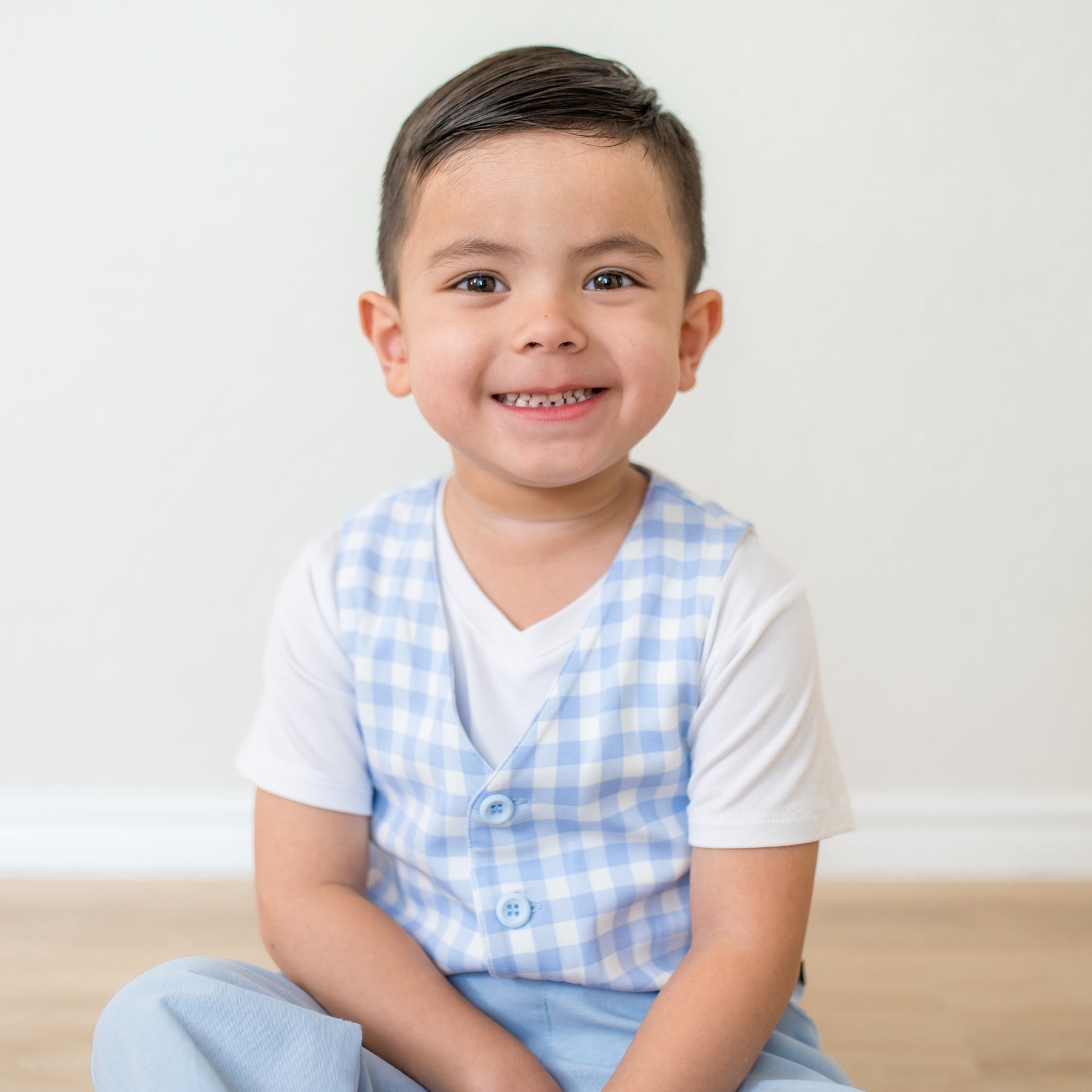 Smiling boy sitting on a wooden floor wearing the Tuxedo Vest in Gingham Breeze overtop a white v-neck
