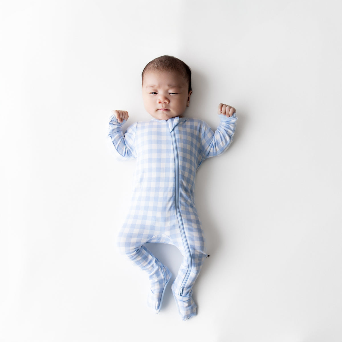 Infant laying on a white surface wearing the Zippered Footie in Gingham Breeze