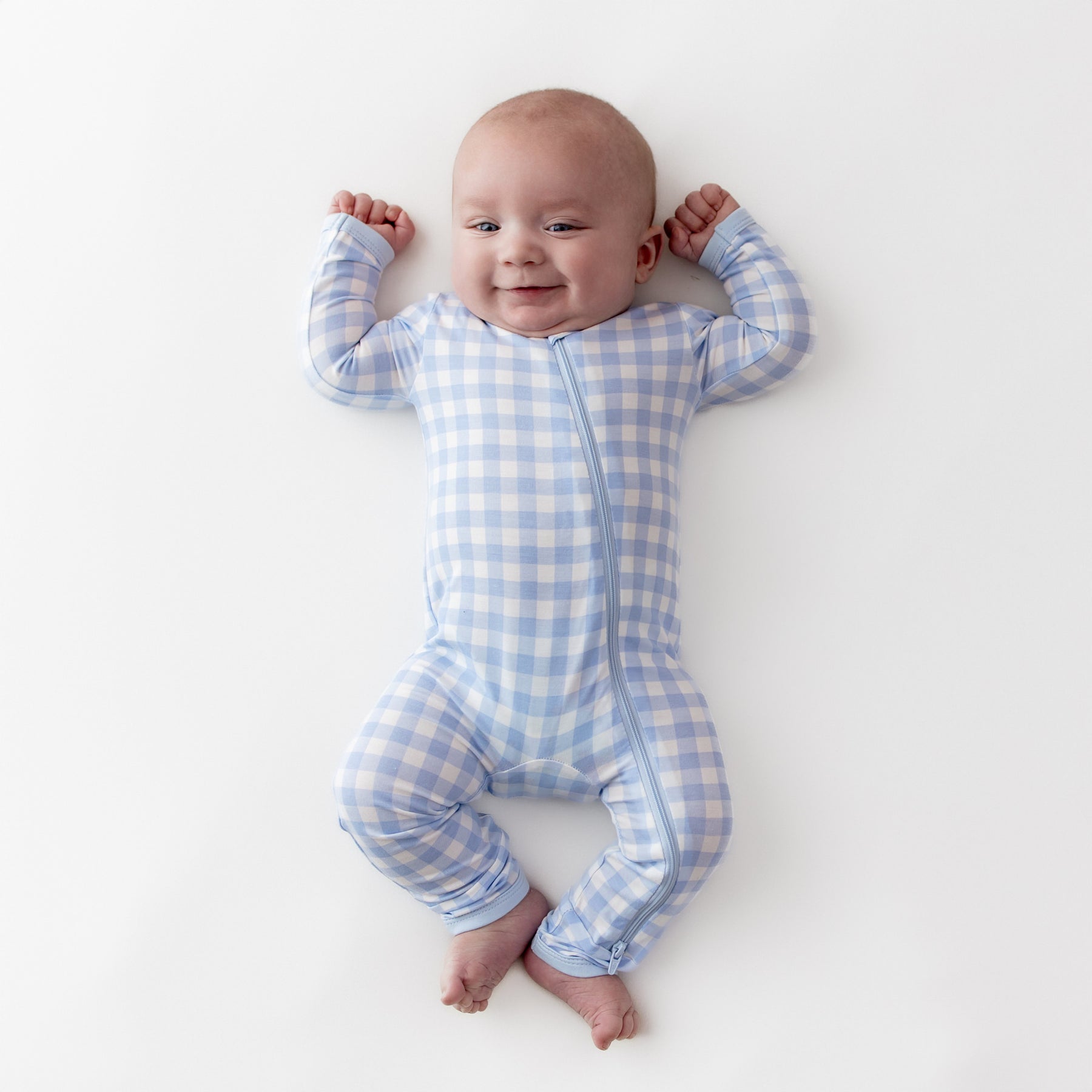 Infant laying on a white surface stretching out wearing the Zippered Romper in Gingham Breeze