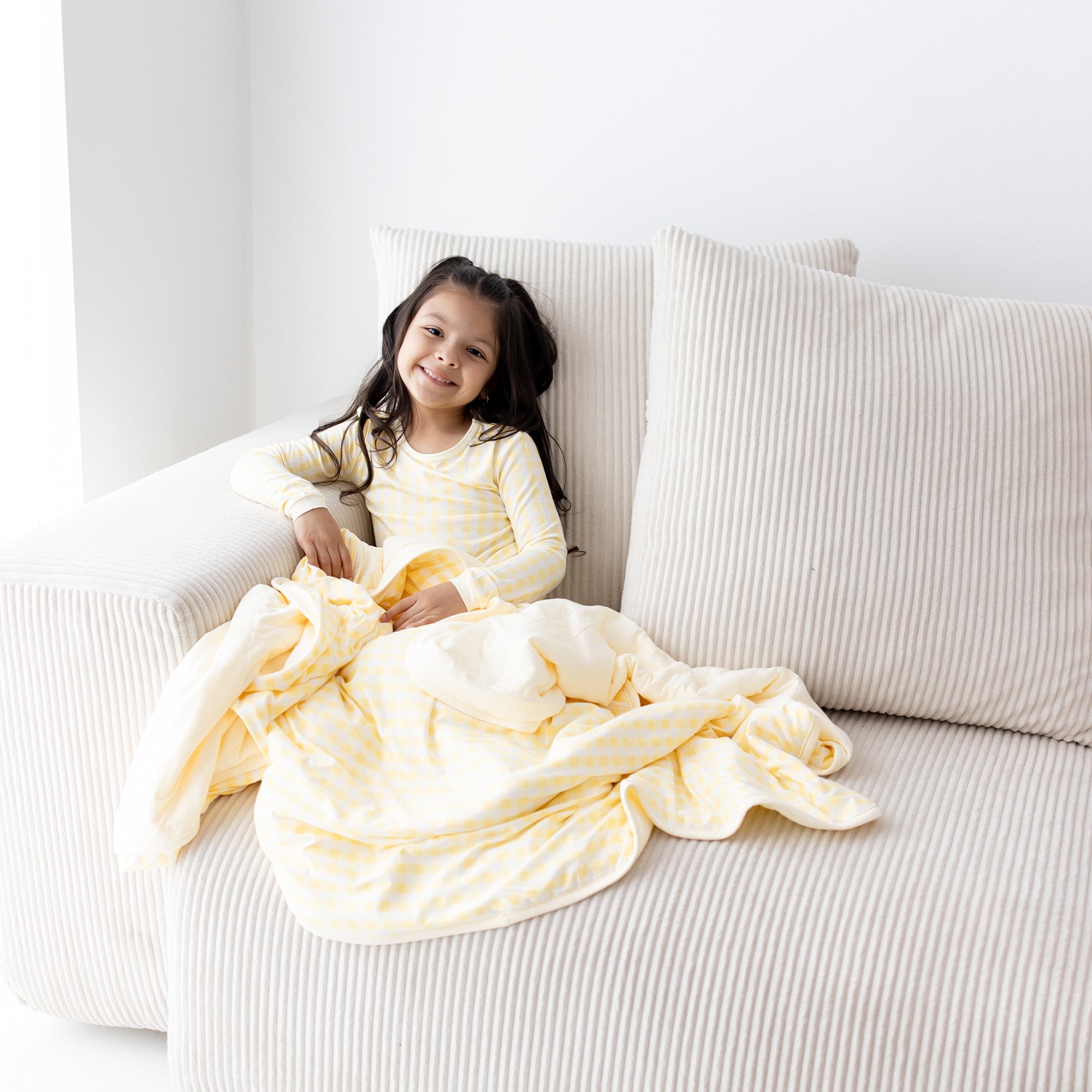 Young girl sitting on the couch with the Toddler Blanket in Gingham Chamomile 1.0 on her lap wearing matching long sleeve pajamas