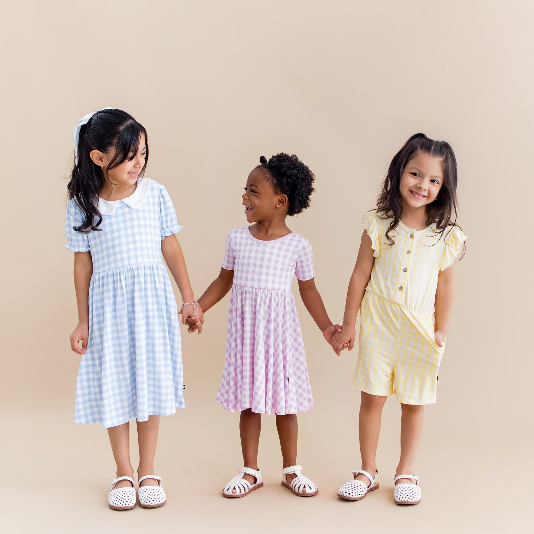 Three girls standing beside one another holding hands wearing the Peter Pan Collar Dress in Gingham Breeze, Twirl Dress in Gingham Thistle and Flutter Sleeve in Gingham Chamomile