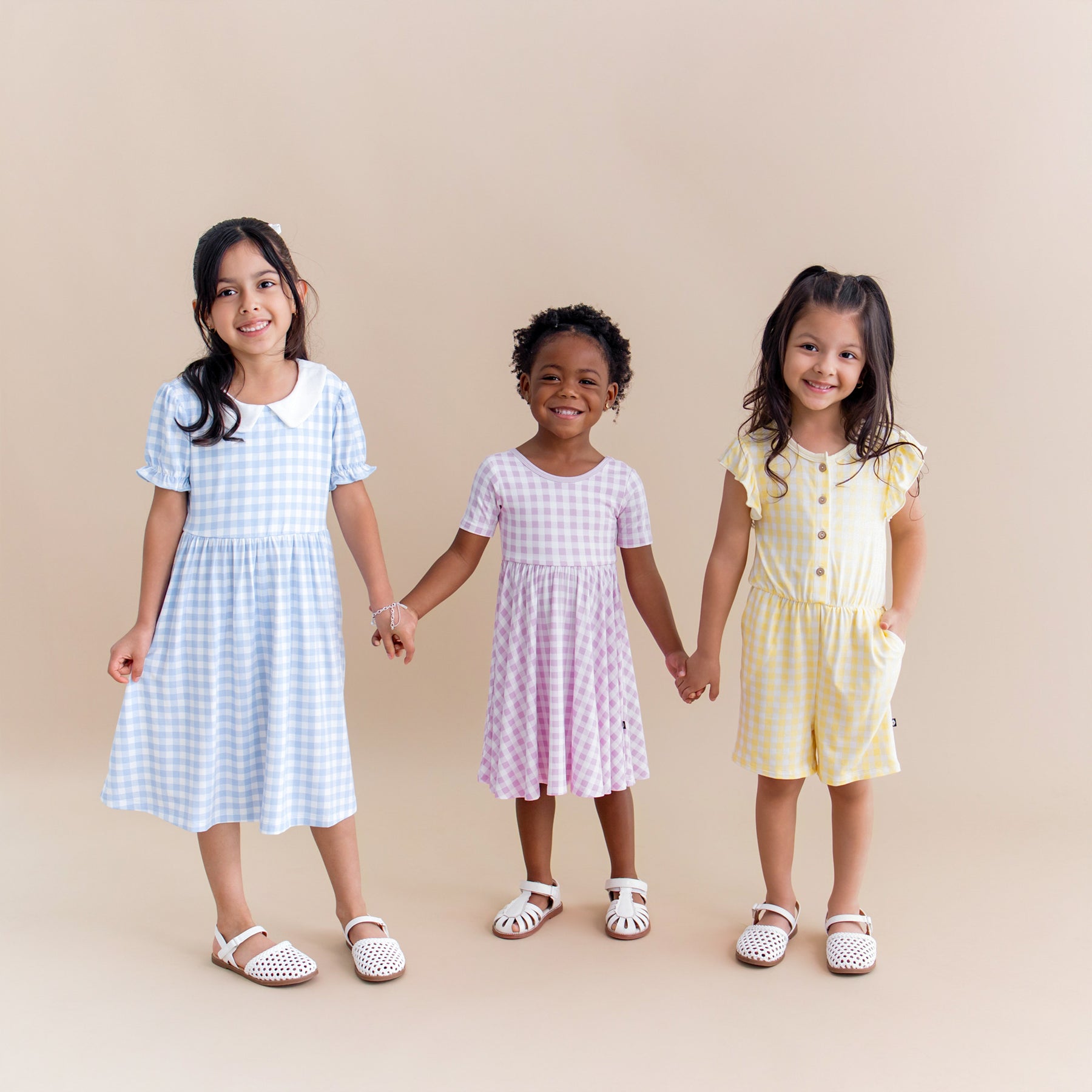 Three young girls standing side by side holding hands wearing the Peter Pan Collar Dress in Gingham Breeze, Twirl Dress in Gingham Thistle and Flutter Sleeve Romper in Gingham Chamomile