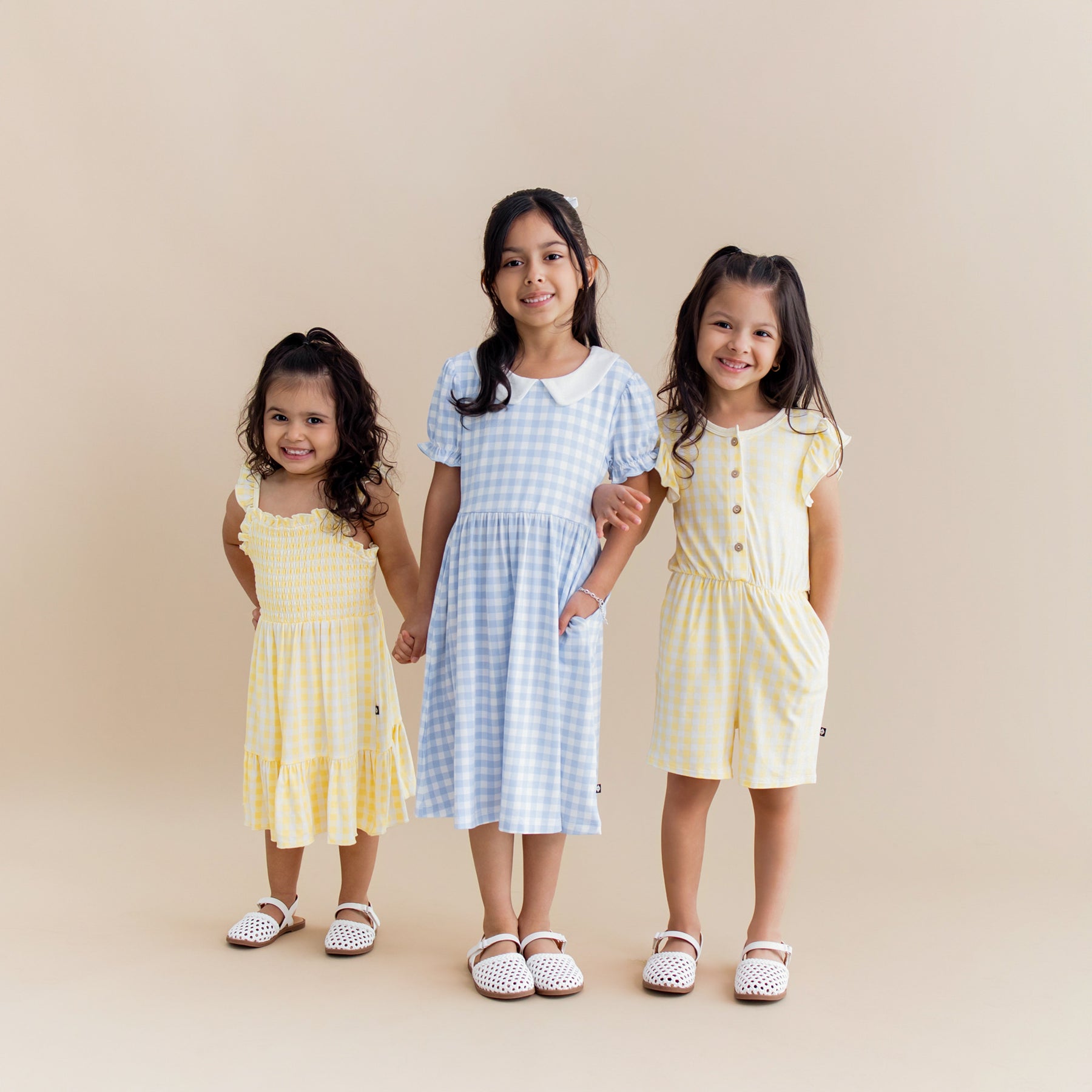 Three young girls standing side by side wearing the Smocked Dress in Gingham Chamomile, Peter pan dress in Gingham Breeze and Flutter sleeve romper in Gingham Chamomile