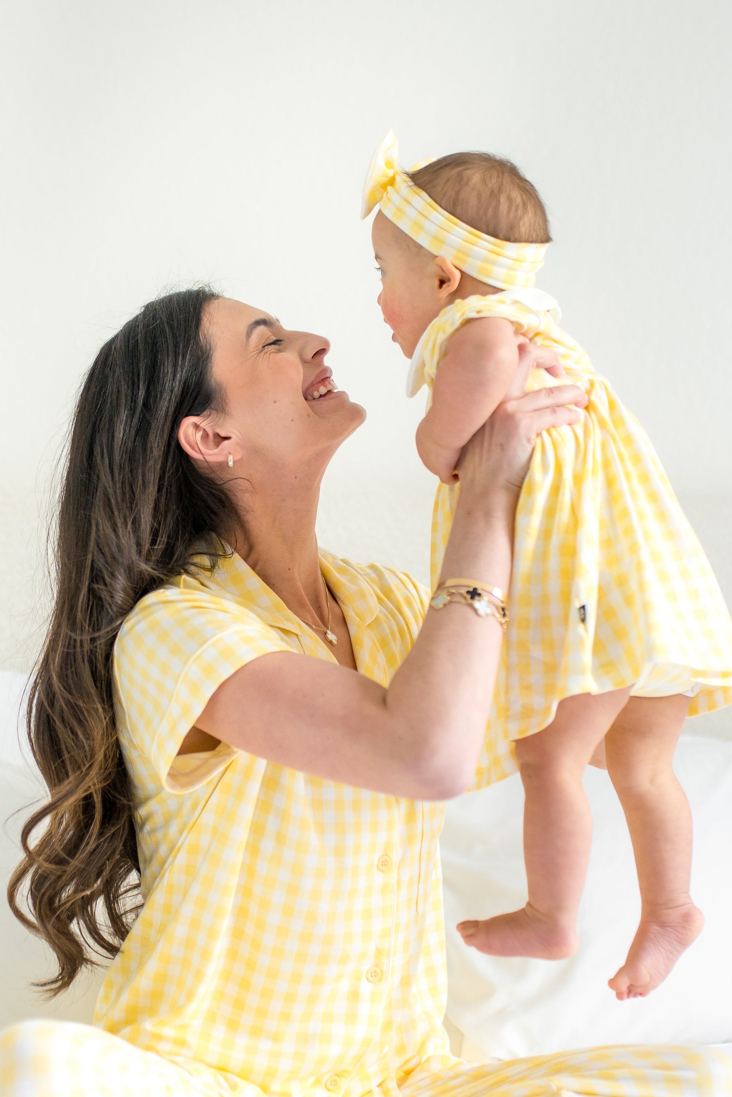 Mother sitting on a bed wearing the Women's Short Sleeve Cropped Pajama Set in Gingham Chamomile holding her young daughter who is wearing a matching peter pan collar bodysuit dress