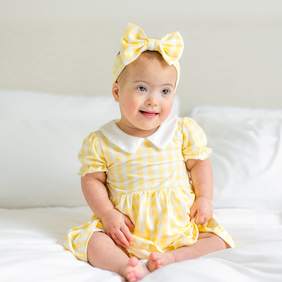 Young infant sitting on a bed wearing the Peter Pan Collar Bodysuit Dress in Gingham Chamomile with matching bow headband