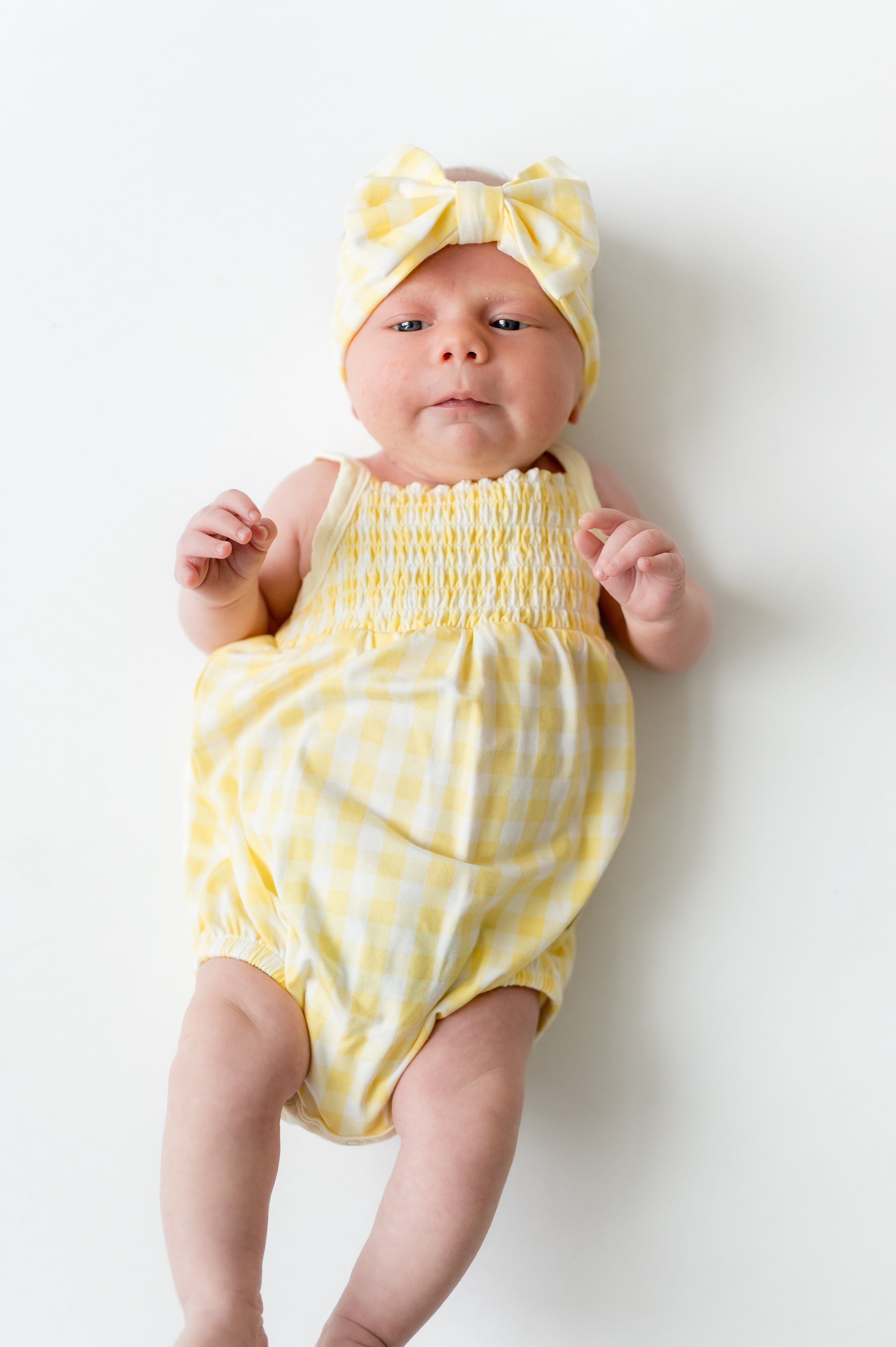 Newborn laying on a white surface wearing the Smocked Bubble Romper in Gingham Chamomile with matching bow