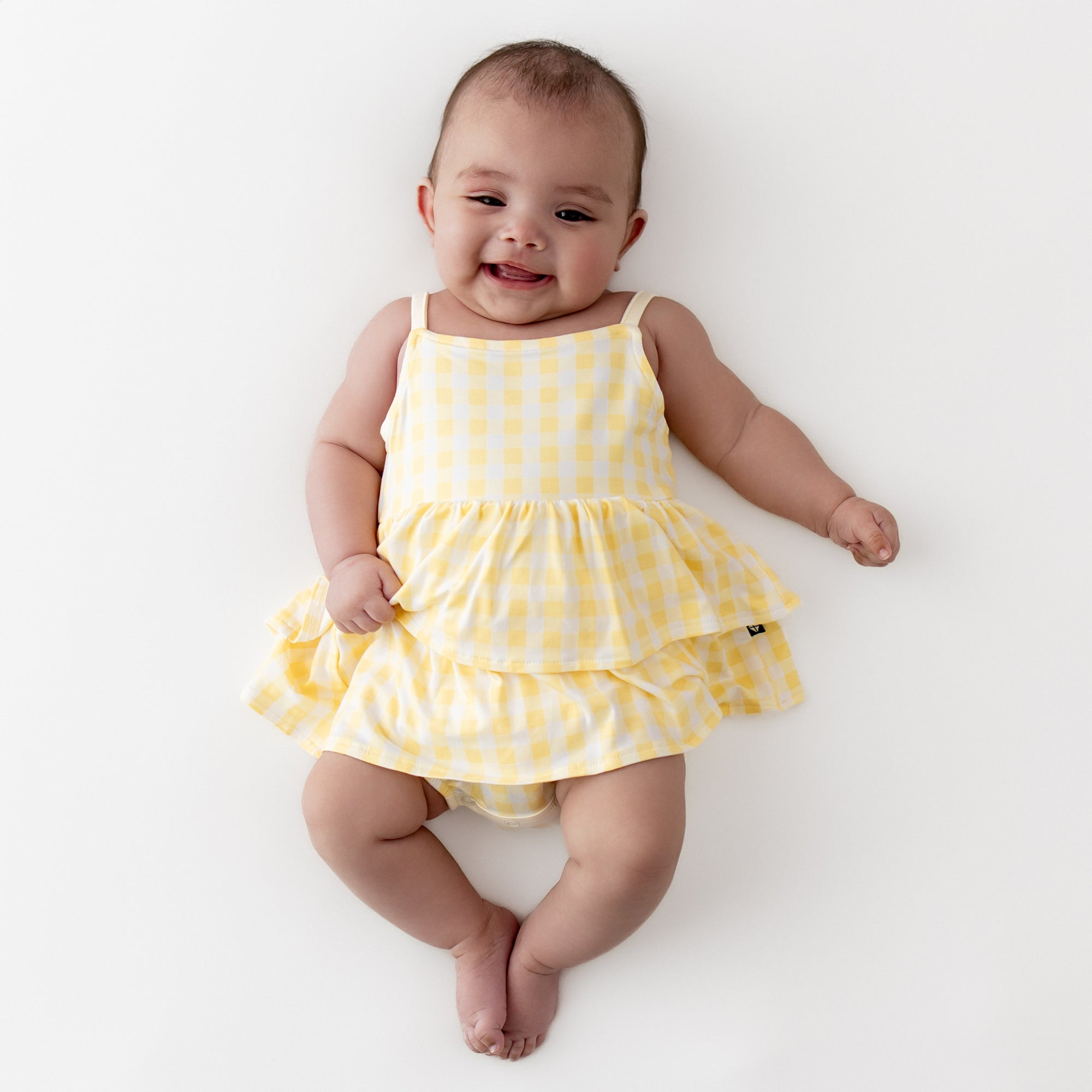 Infant girl wearing the Spaghetti Strap Ruffle Romper in Gingham Chamomile laying on a light beige surface