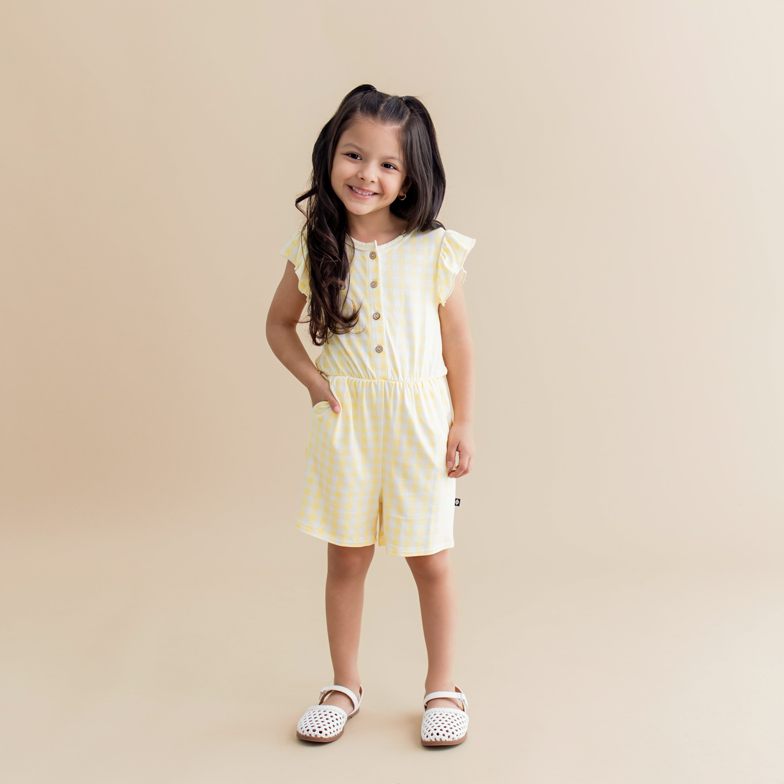 Young smiling girl standing with her hand in the pocket of the Flutter Sleeve Romper in Gingham Chamomile
