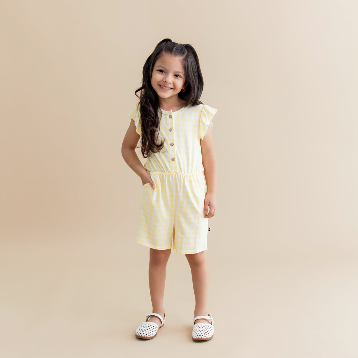 Young smiling girl standing with her hand in the pocket of the Flutter Sleeve Romper in Gingham Chamomile