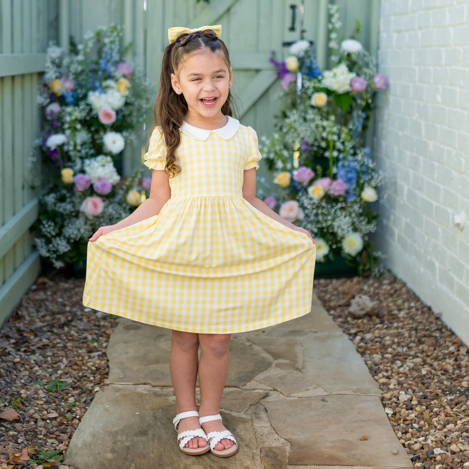 Young girl standing on a stone path in front of flowers and a green fence wearing the Peter Pan Collar Dress in Gingham Chamomile holding out the skirt