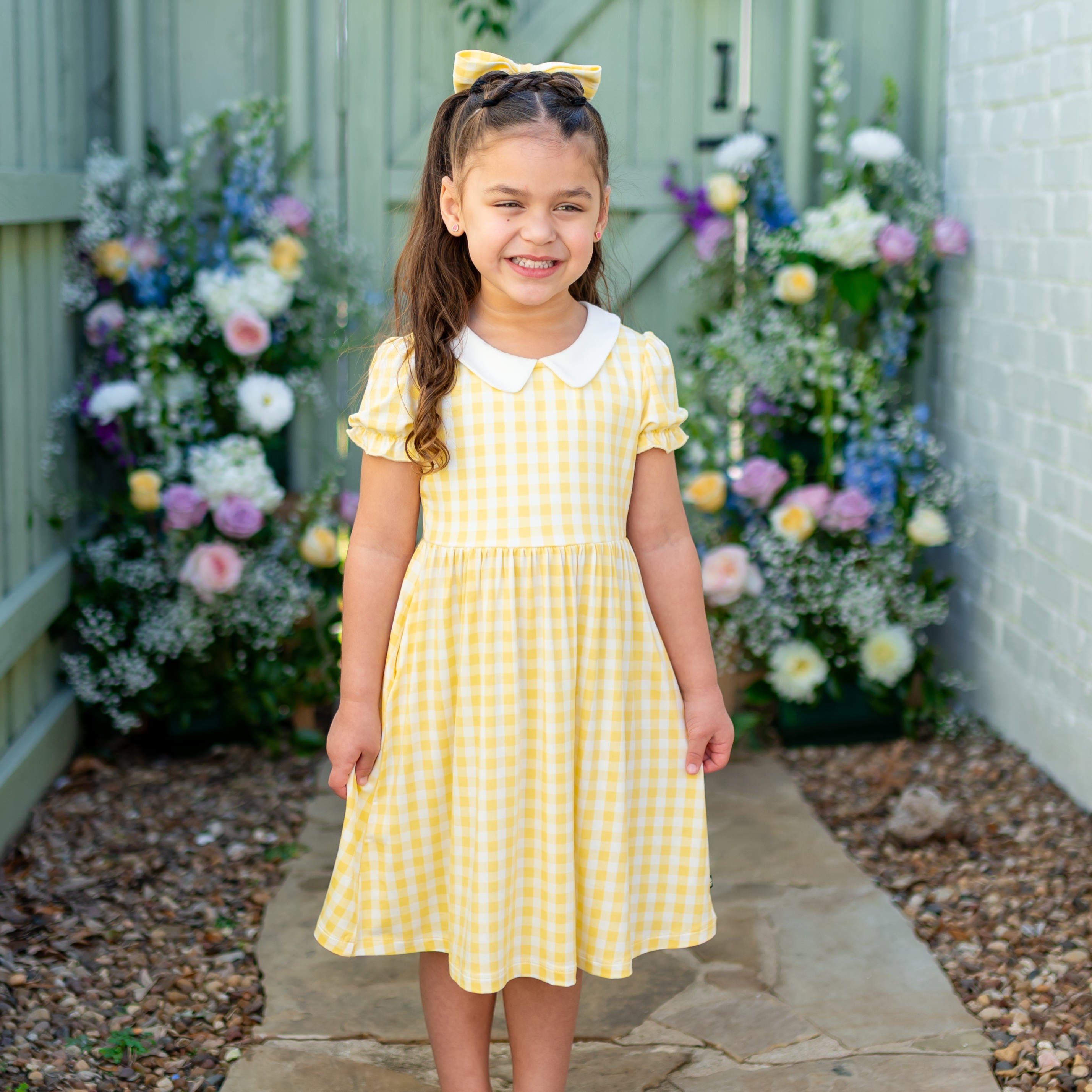 Smiling girl standing on a stone concrete in front of flowers wearing the Peter Pan Collar Dress in Gingham Chamomile