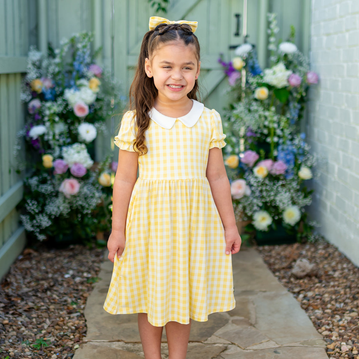 Smiling girl standing on a stone concrete in front of flowers wearing the Peter Pan Collar Dress in Gingham Chamomile