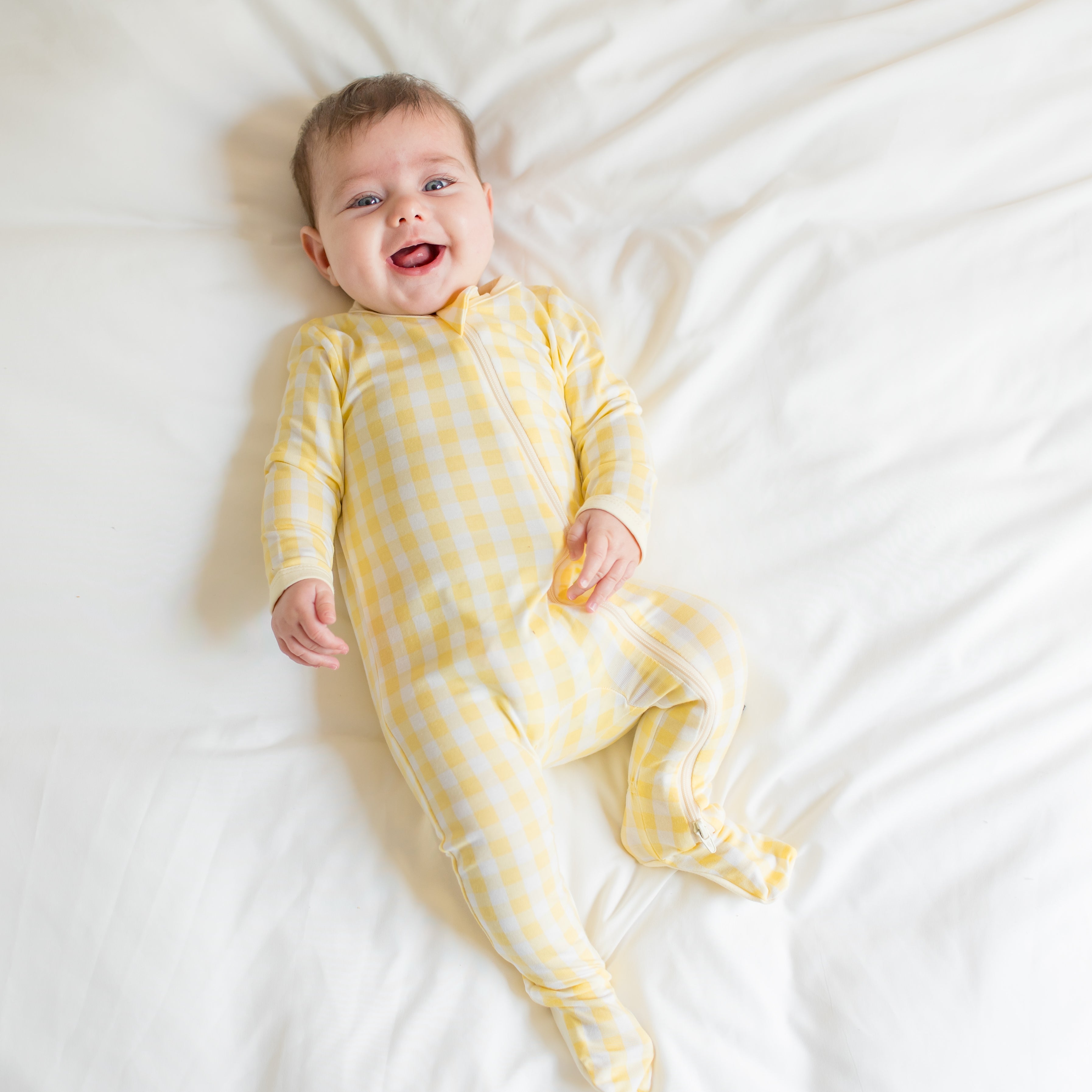 Young infant laying in a bed wearing the Zippered Footie in Gingham Chamomile