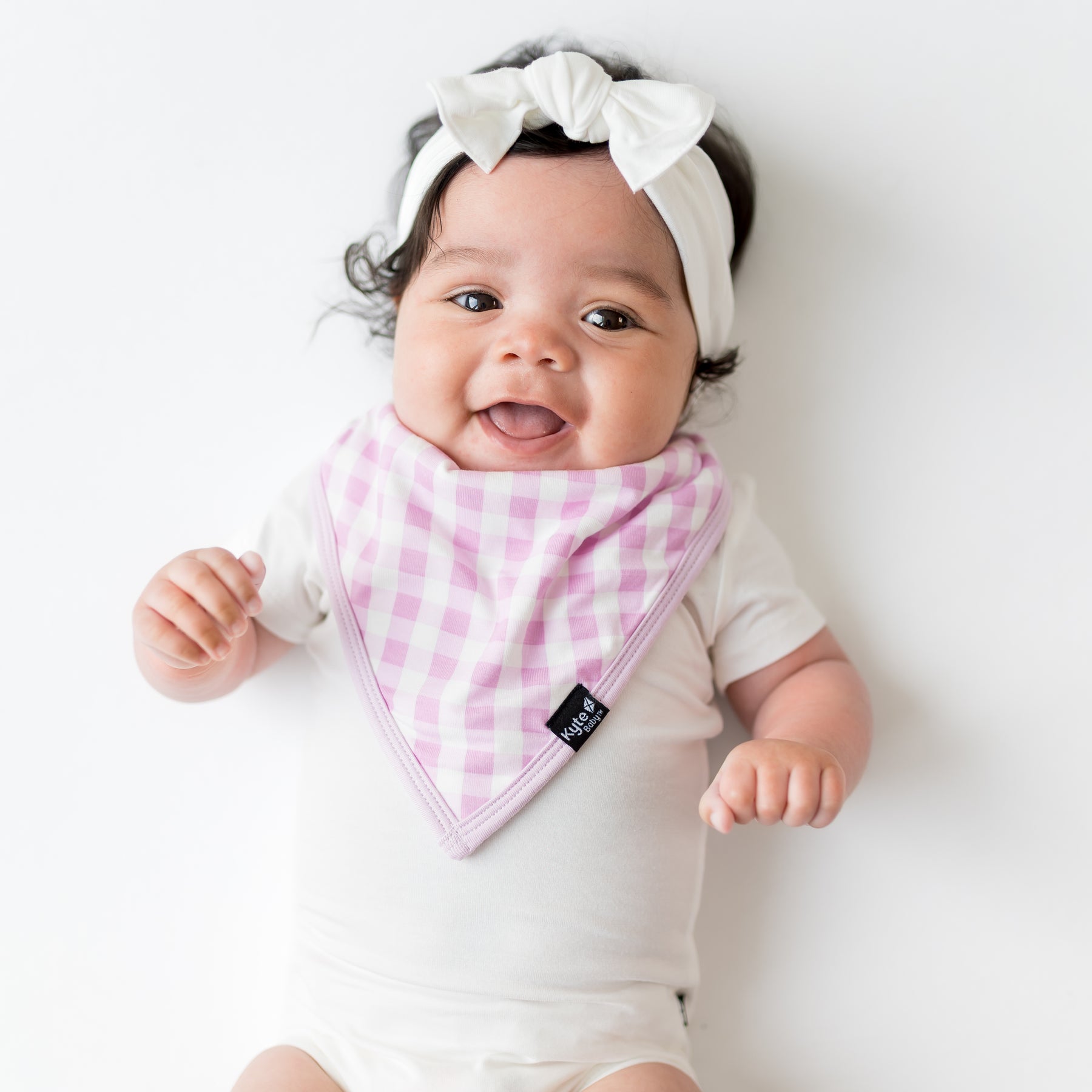Infant girl wearing the Bib in Gingham Thistle overtop a white bodysuit and white bow