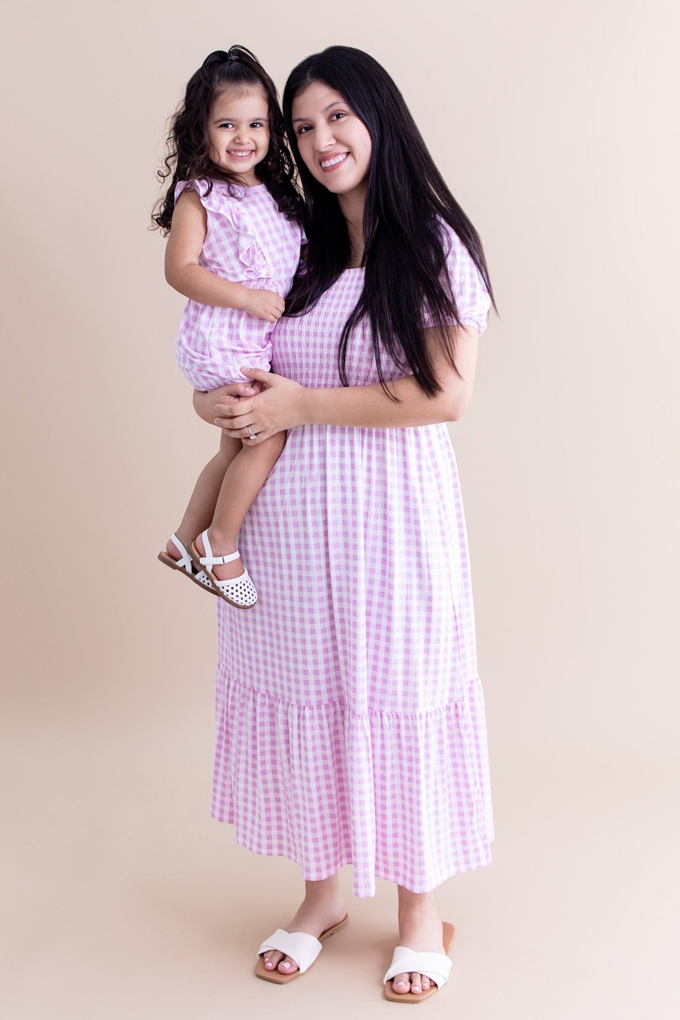 Smiling female model wearing the Women’s Smocked Dress in Gingham Thistle holding her daughter who is matching in a bubble romper