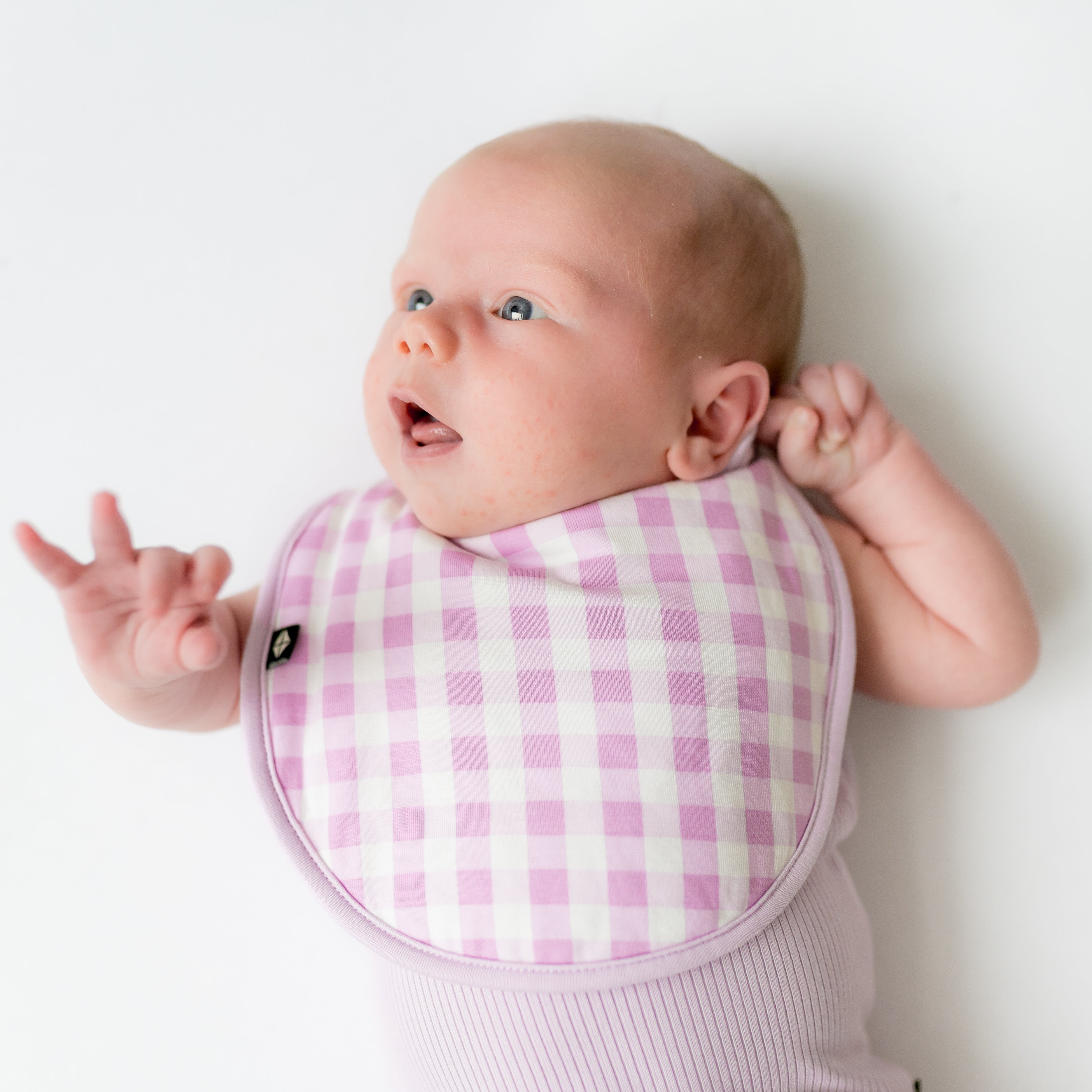 Newborn baby laying on a white surface wearing the Cushy Bib in Gingham Thistle