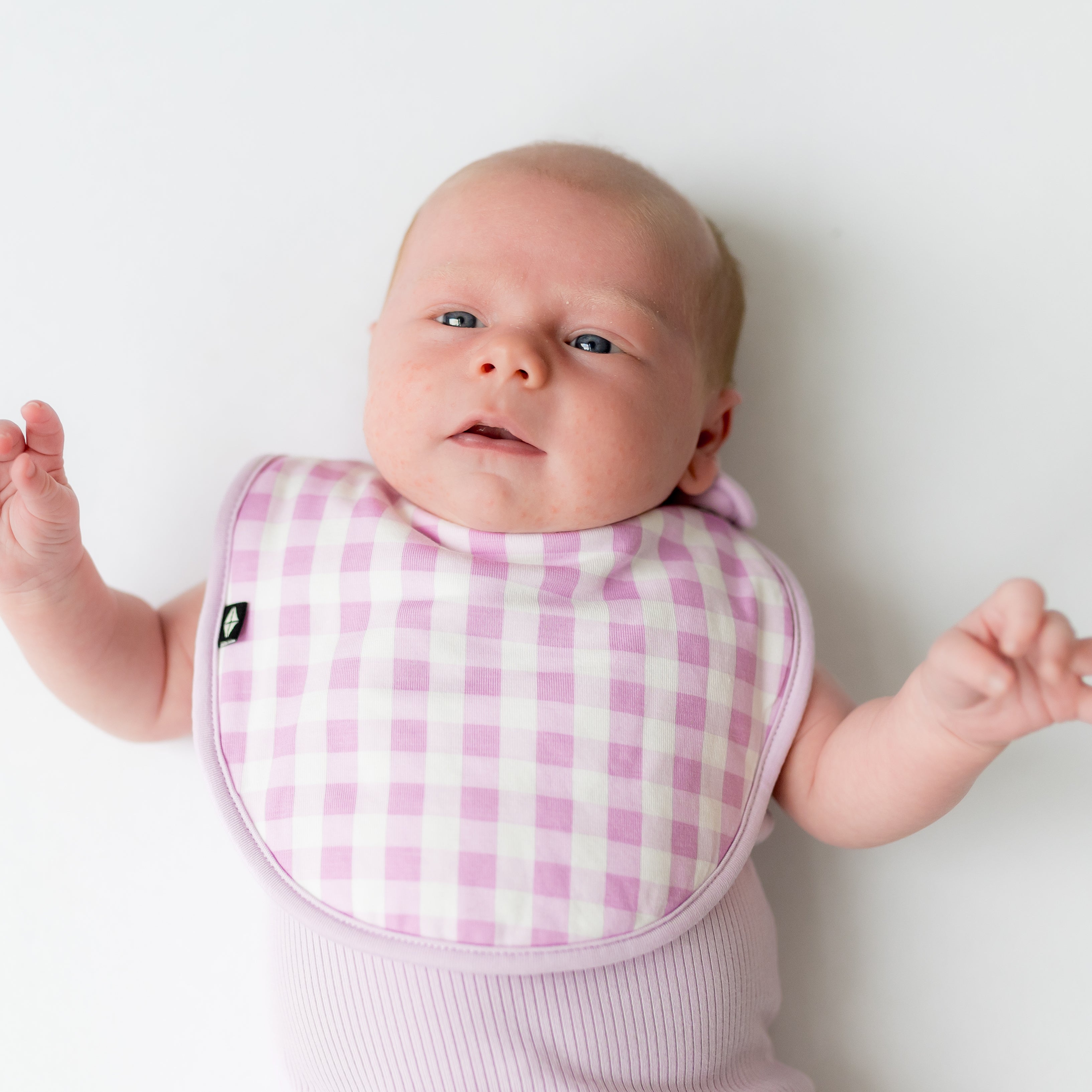 Newborn laying on a white surface wearing the Cushy Bib in Gingham Thistle