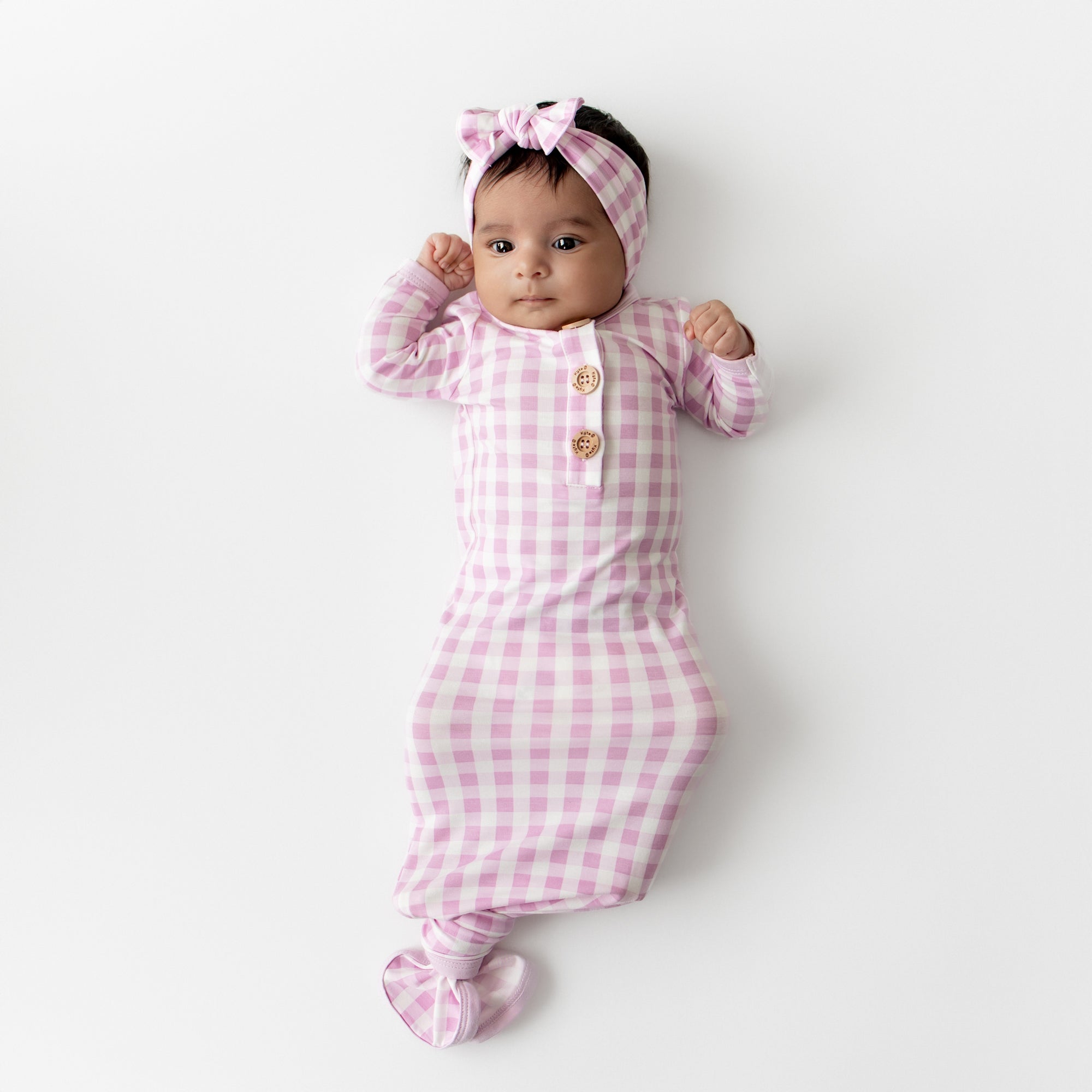 Infant girl laying on a white surface wearing the Knotted Gown with Bow Set in Gingham Thistle