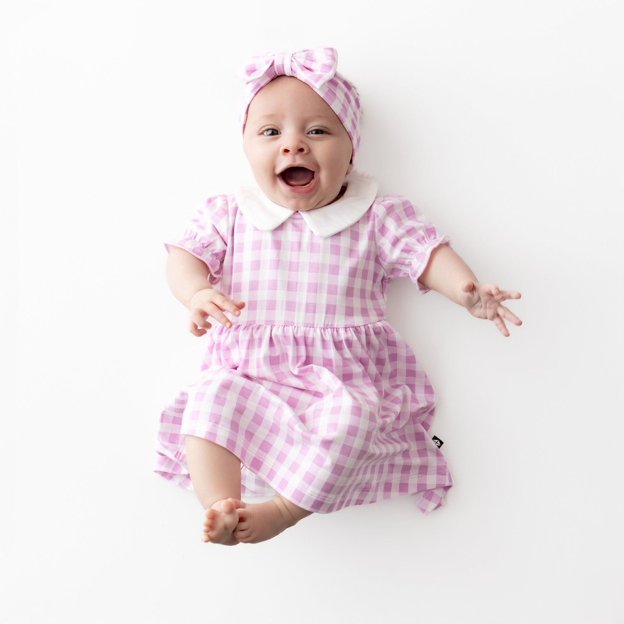 Smiling infant laying on a white surface wearing the Peter Pan Collar Bodysuit Dress in Gingham Thistle