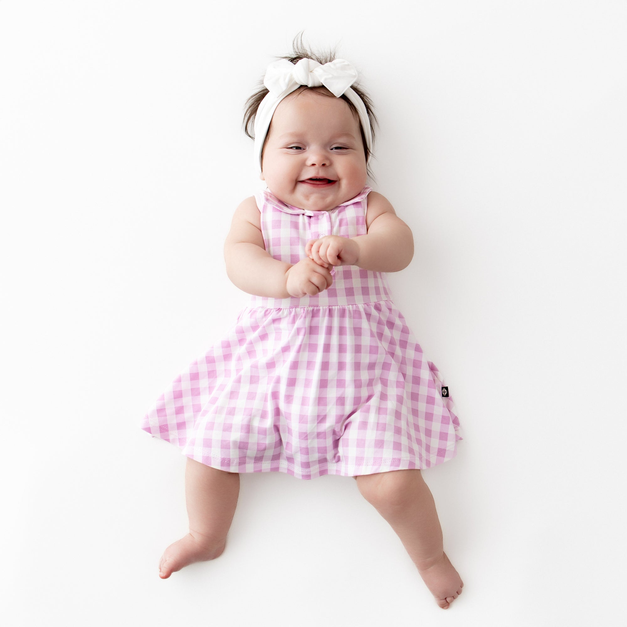 Smiling infant wearing the Polo Bodysuit Dress in Gingham Thistle with a white knotted bow headband on a light cream surface