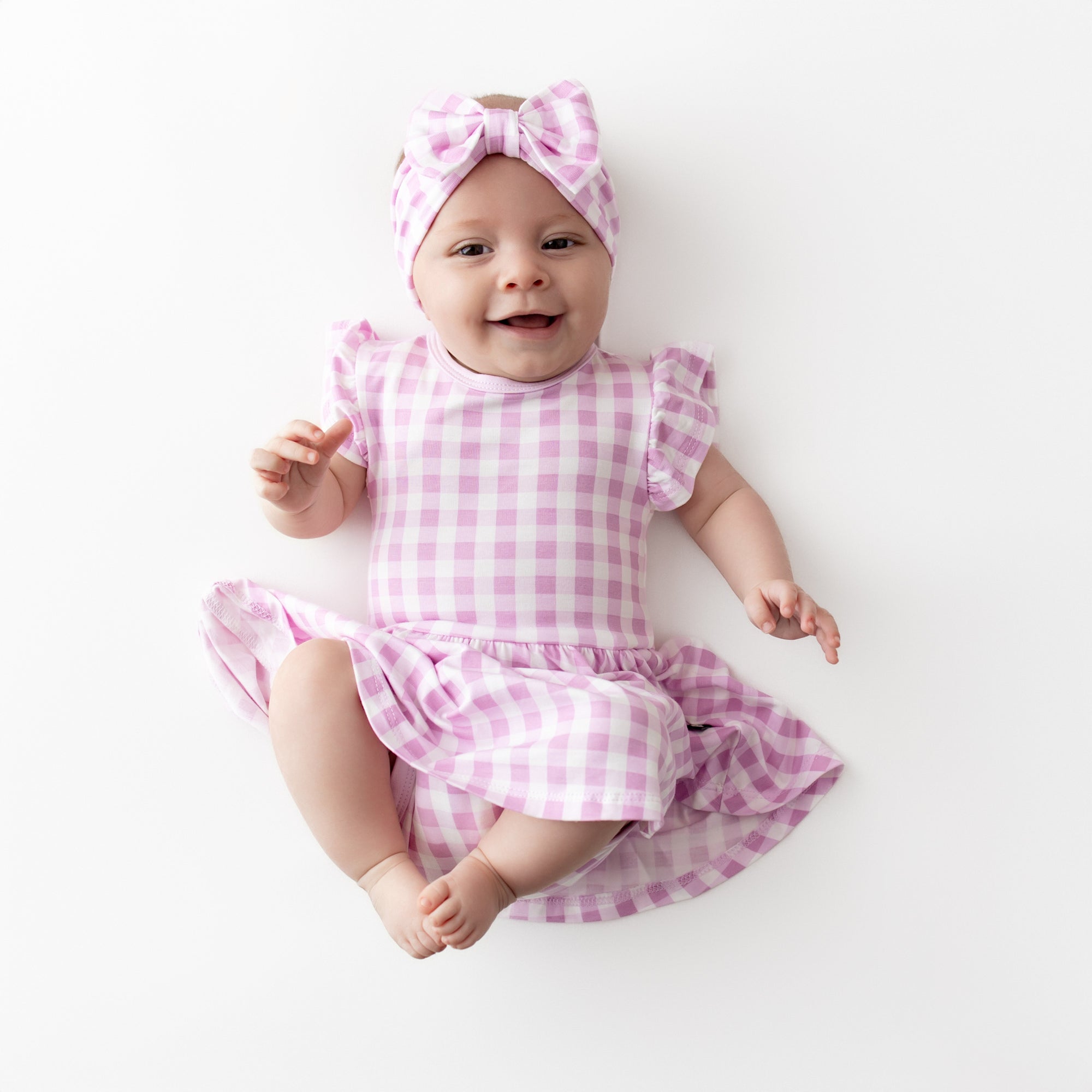 Smiling infant wearing the Twirl Bodysuit Dress in Gingham Thistle with matching bow headband