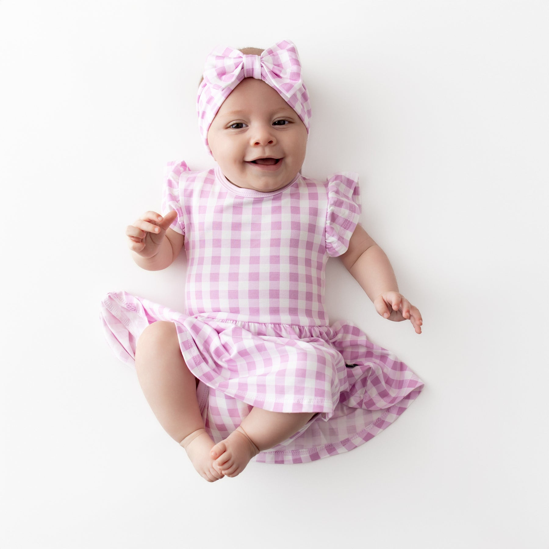 Smiling infant wearing the Twirl Bodysuit Dress in Gingham Thistle with matching bow headband