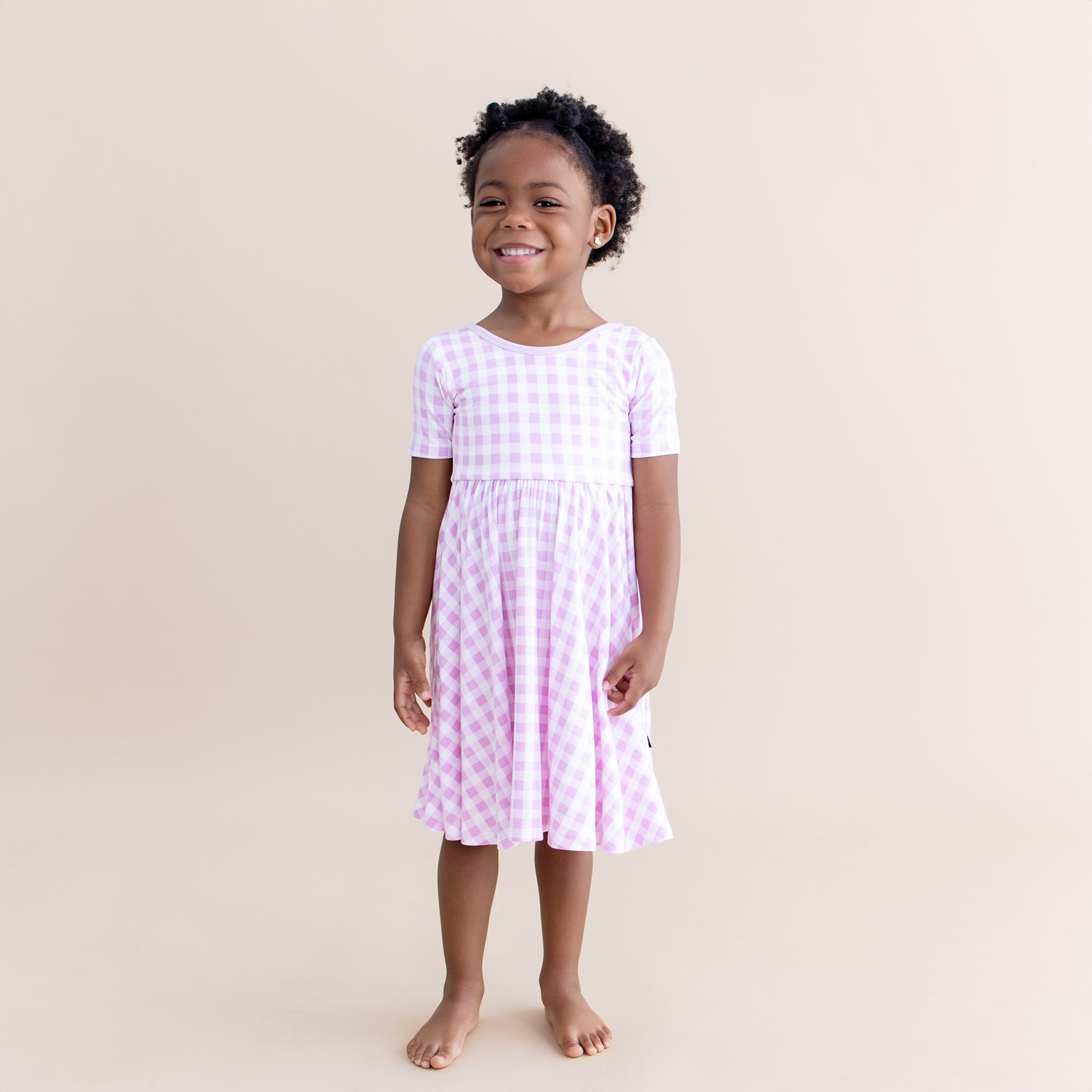 Young girl smiling wearing the Twirl Dress in Gingham Thistle standing in front of a light beige background