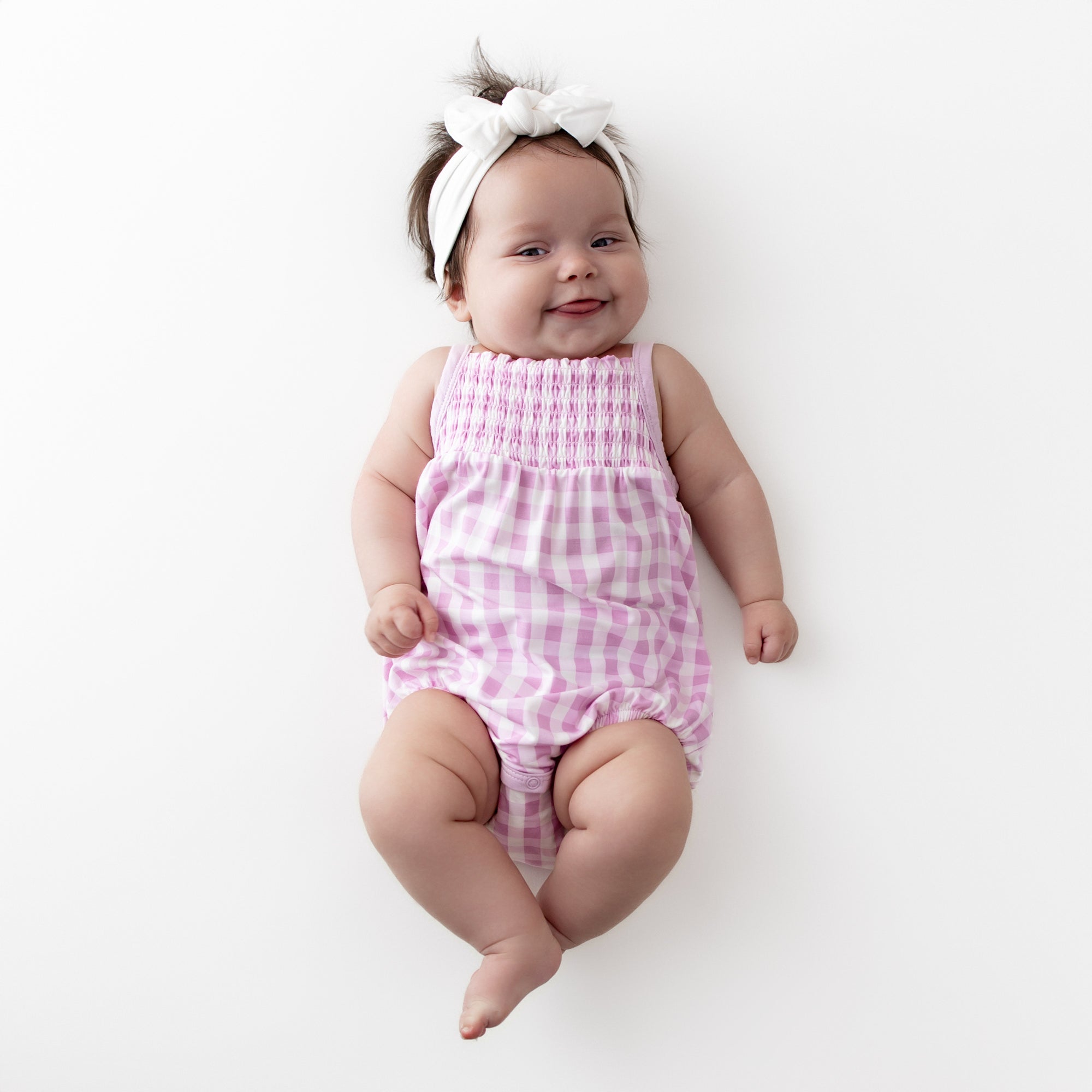 Smiling infant wearing the Smocked Bubble Romper in Gingham Thistle with a white knotted bow headband