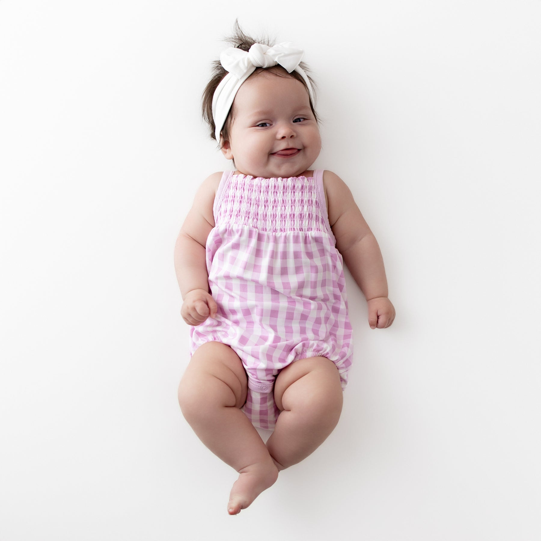 Smiling infant wearing the Smocked Bubble Romper in Gingham Thistle with a white knotted bow headband